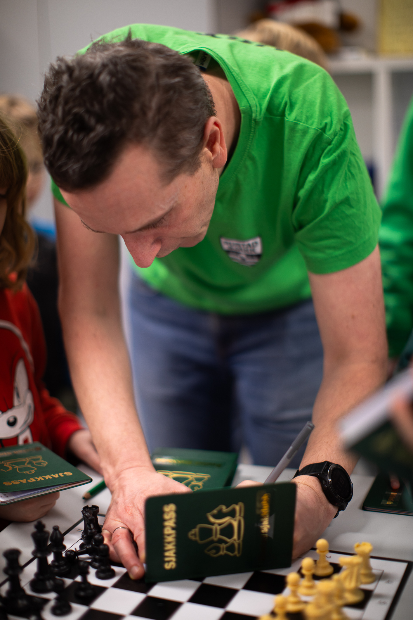 Mann står bøyd over et sjakkbrett med en liten blå bok med gullskrift på hvor det står Sjakkpass utenpå.