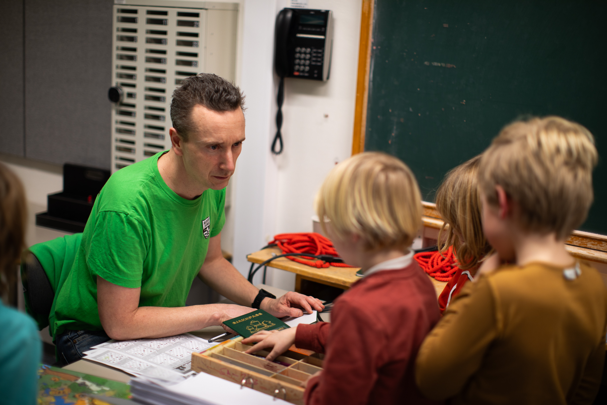 Mann i grønn T-skjorte snakker med en liten rekke barn i småskolealder. De står på den andre siden av bordet der han sitter.