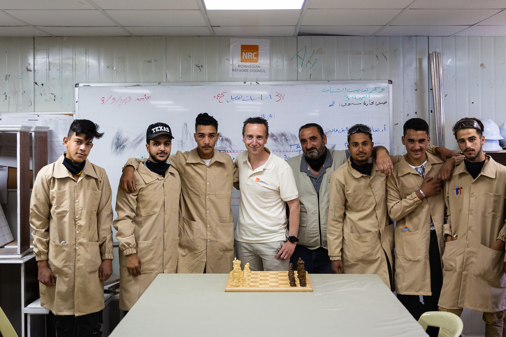 Norsk mann i hvit T-skjorte står sammen med en rekke menn i beige frakker. Fremmed skriftspråk på tavlen bak. På bordet foran dem står et sjakkbrett.