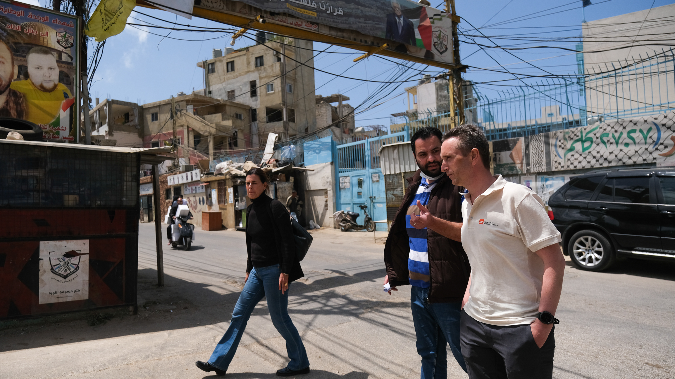 Norsk mann går over gaten i en by i midtøsten sammen med en mann og en kvinne i vestlige klær. EUndt dem er boligblokker i betong og en masse strømledninger i lufta.