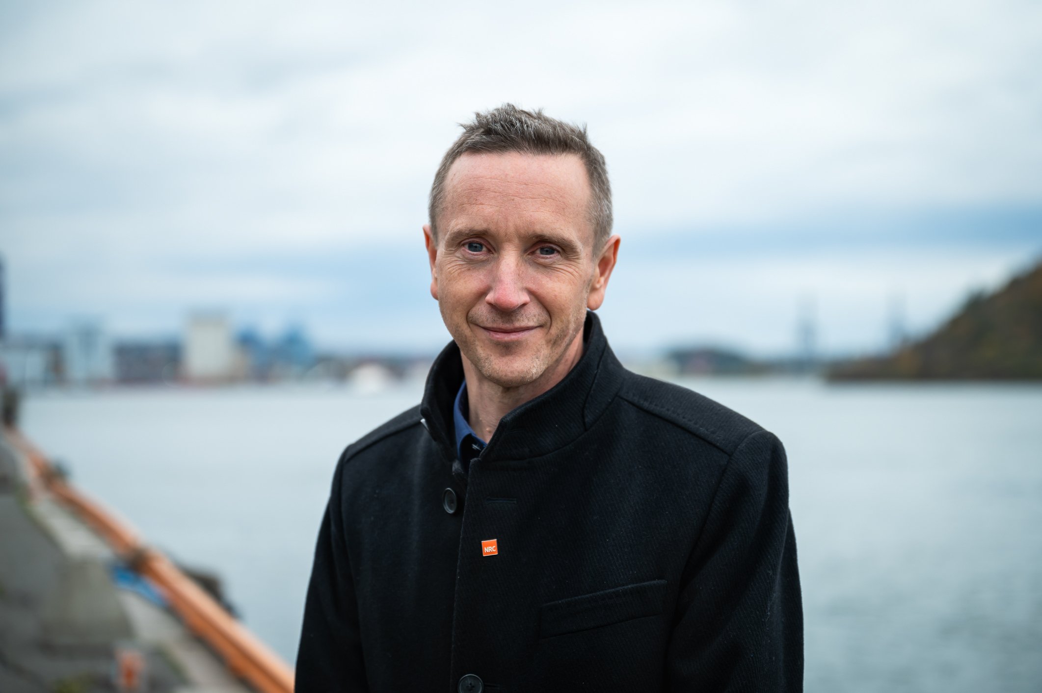 Mann i mørk jakke ser rett i kamera. Står på kaikant med sjø og havn i bakgrunnen. Bildet er tatt fra brystet og opp.