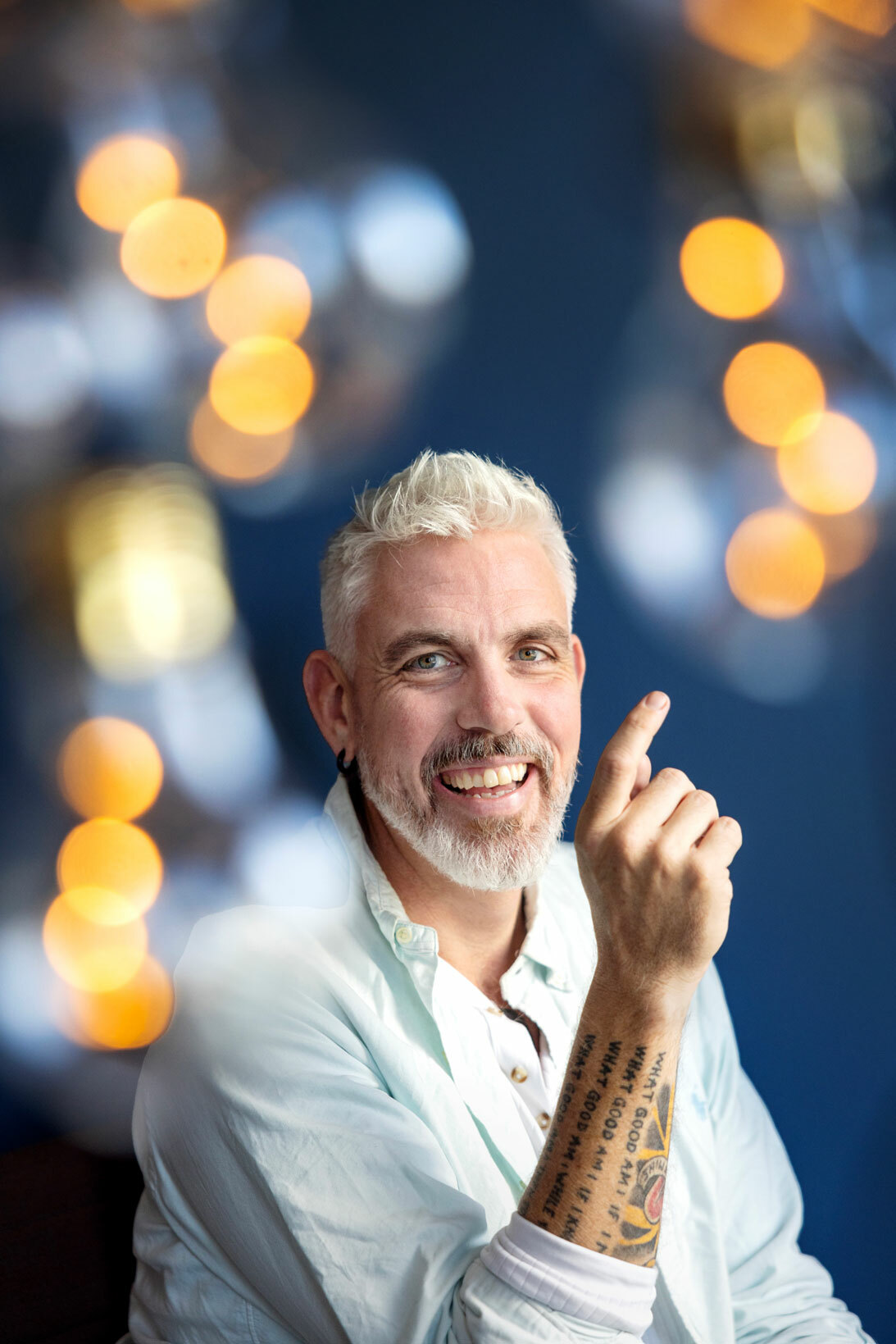 Foto: Mann med grått hår og skjegg sitter og peker med hånden oppover. Han har tatovert inn mye tekst på den.