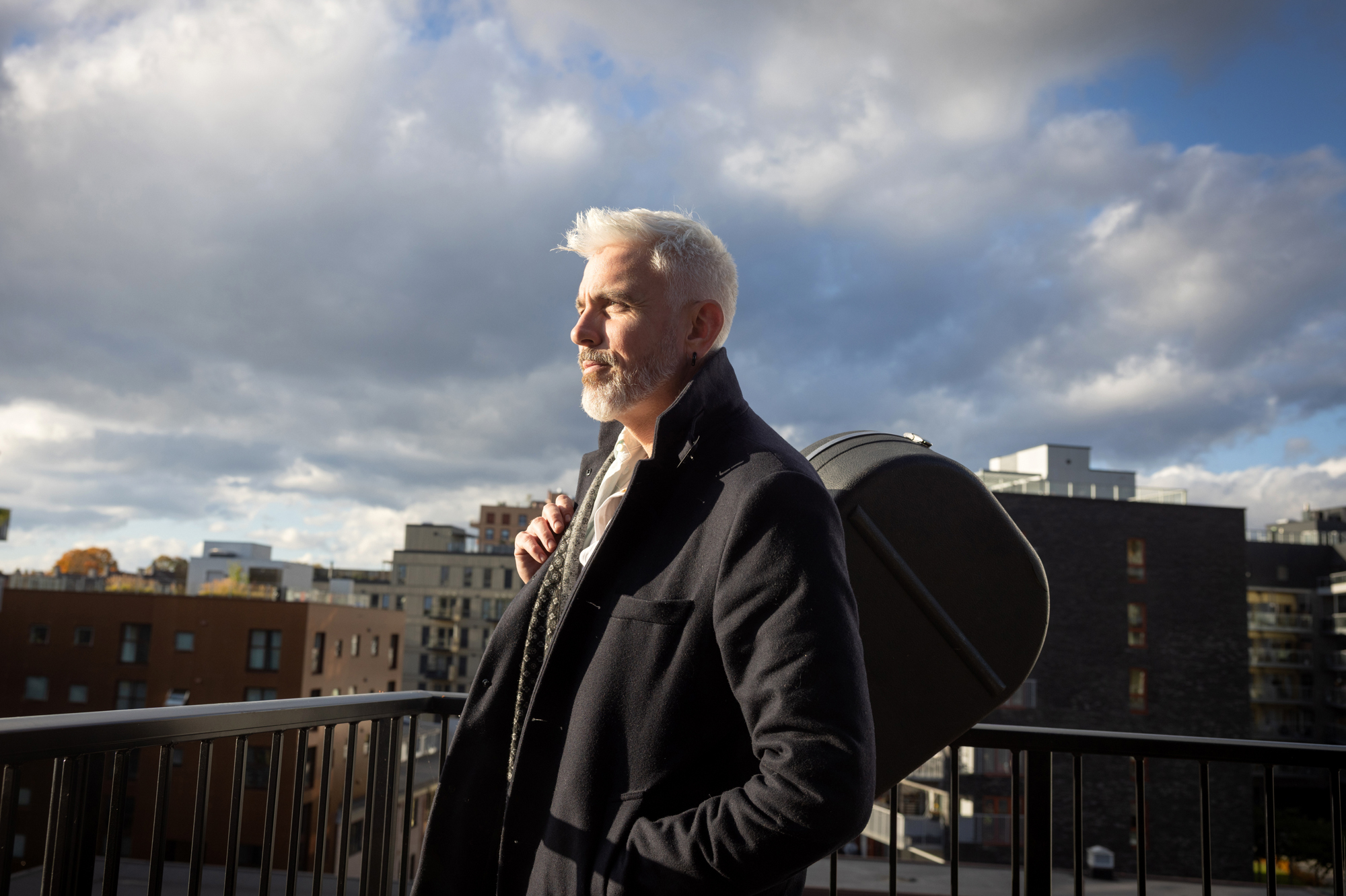 Foto: Mann med grått hår og skjegg har på seg frakk og gitaren over skulderen. Han står på en terrasse og det er skyer på himmelen, men noen solstråler kommer igjennom og det er en flik av blå himmel her og der.