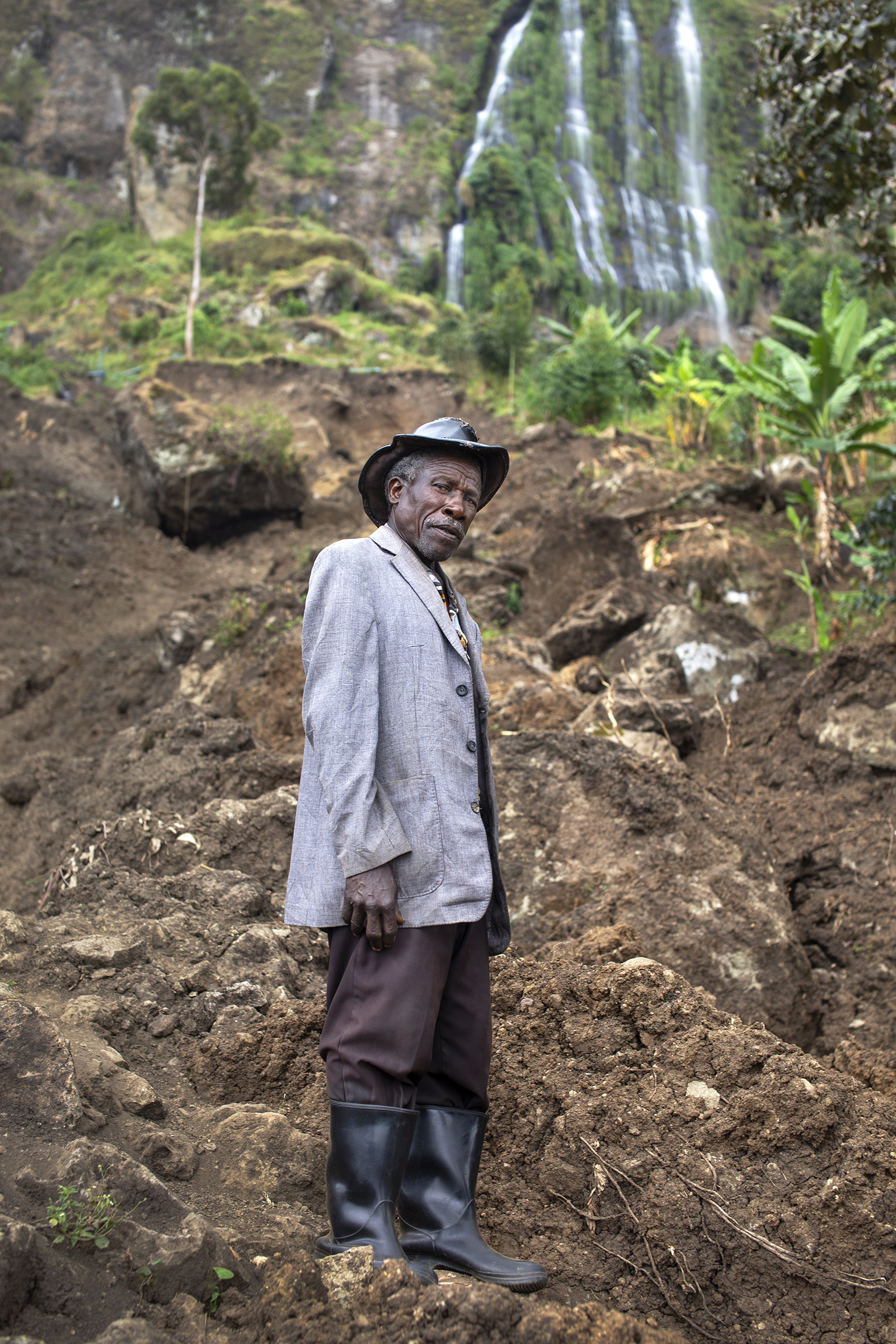 Mr. Akello har på seg lange, sorte støvler og står i jorden som har rast forbi huset hans.  Han har på seg en grå jakke og hatt.