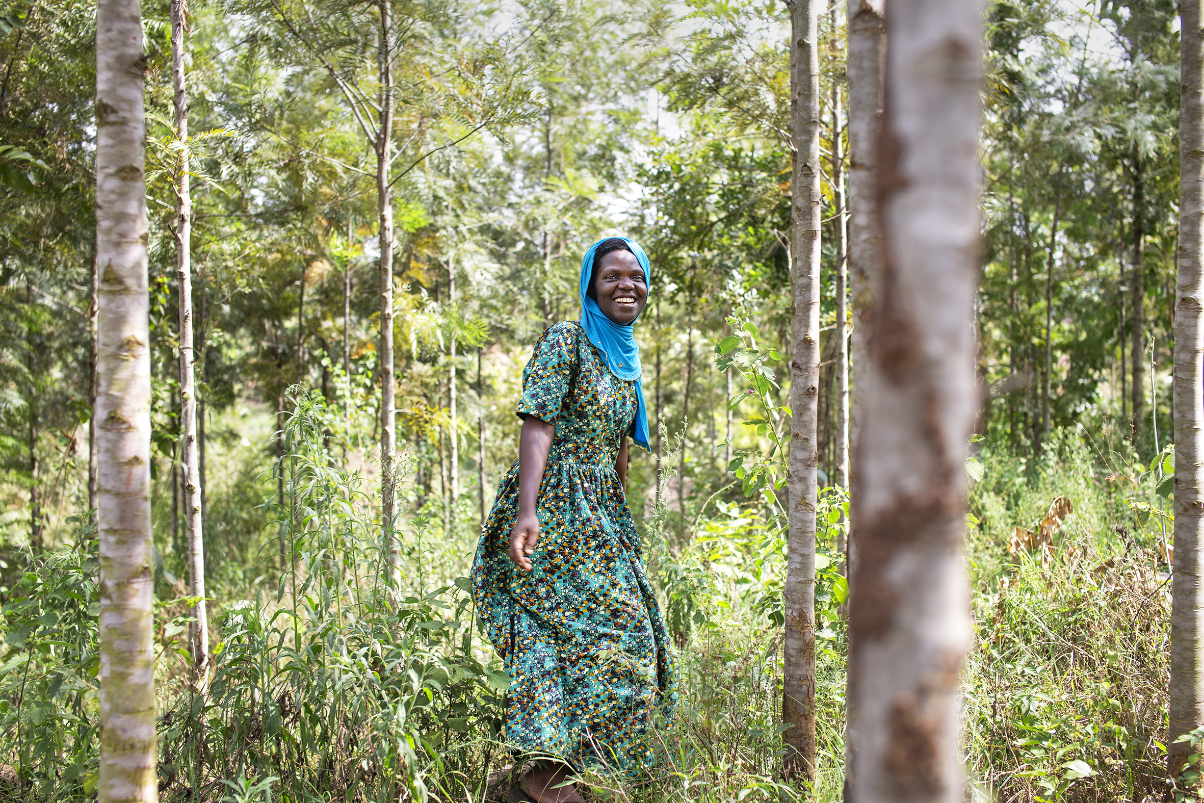 En ung kvinne i fargerik kjole smiler mot kameraet. Hun står i et grønt og frodig landskap.