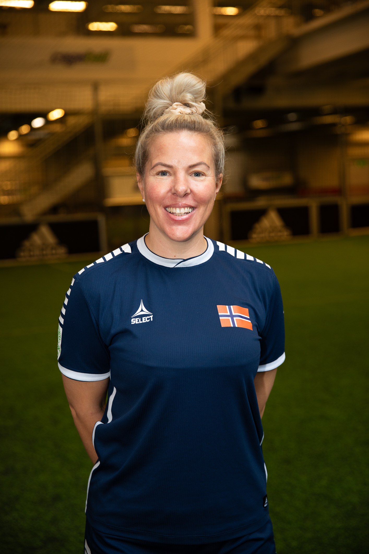 Foto: en kvinne i blå fotballdrakt står og smiler til kamera. I bakgrunnen sees en ballbinge.