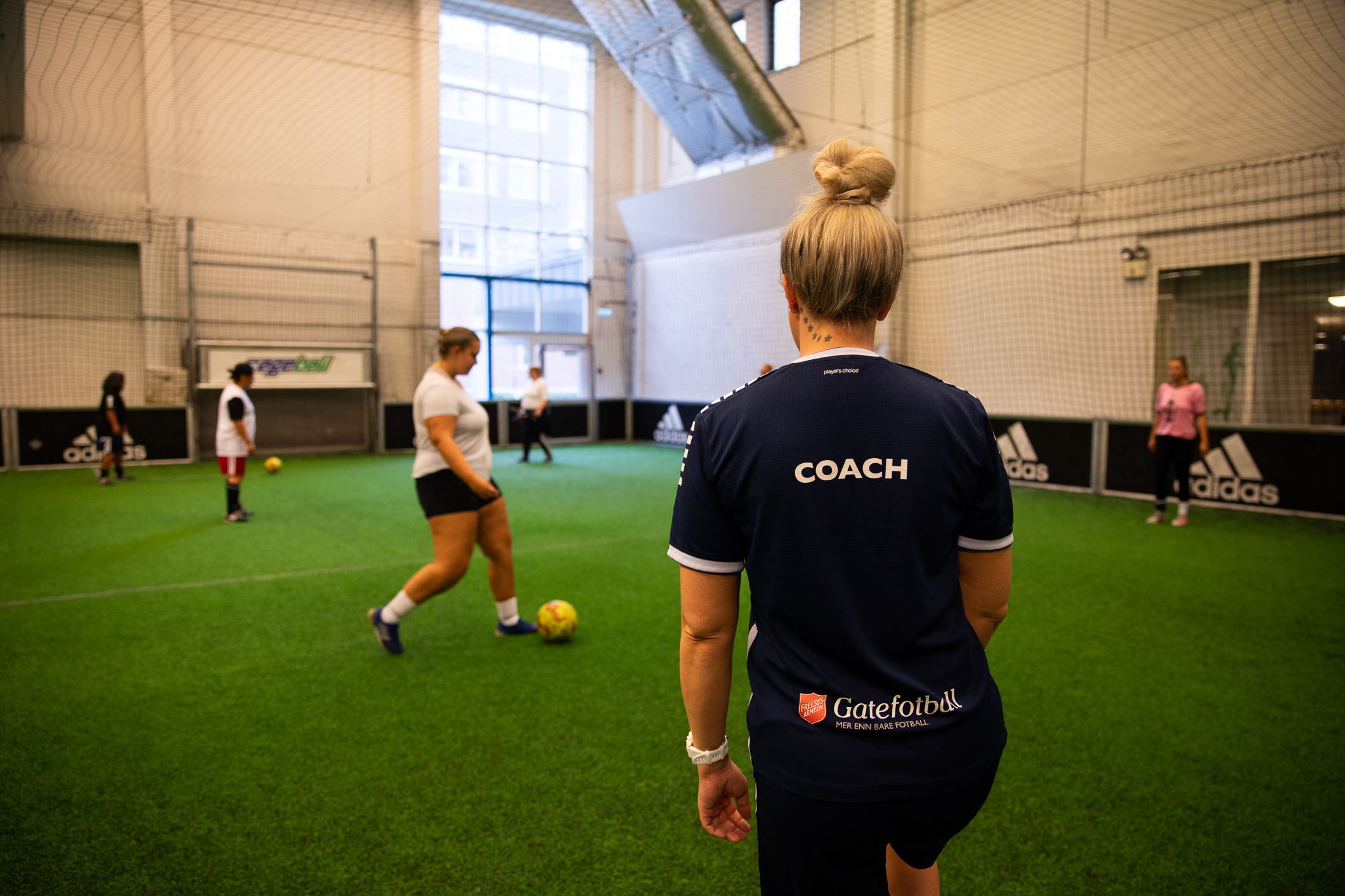Foto: En kvinne står med ryggen mot kamera i blå fotballdrakt med ordet coach på ryggen. Hun ser mot spillere som varmer opp i ballbingen.