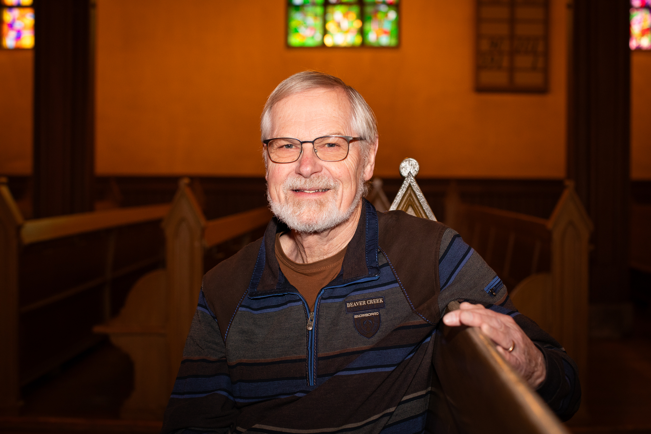 Gammel mann med briller og mørke klær sitter på en kirkebenk i et stort rom.