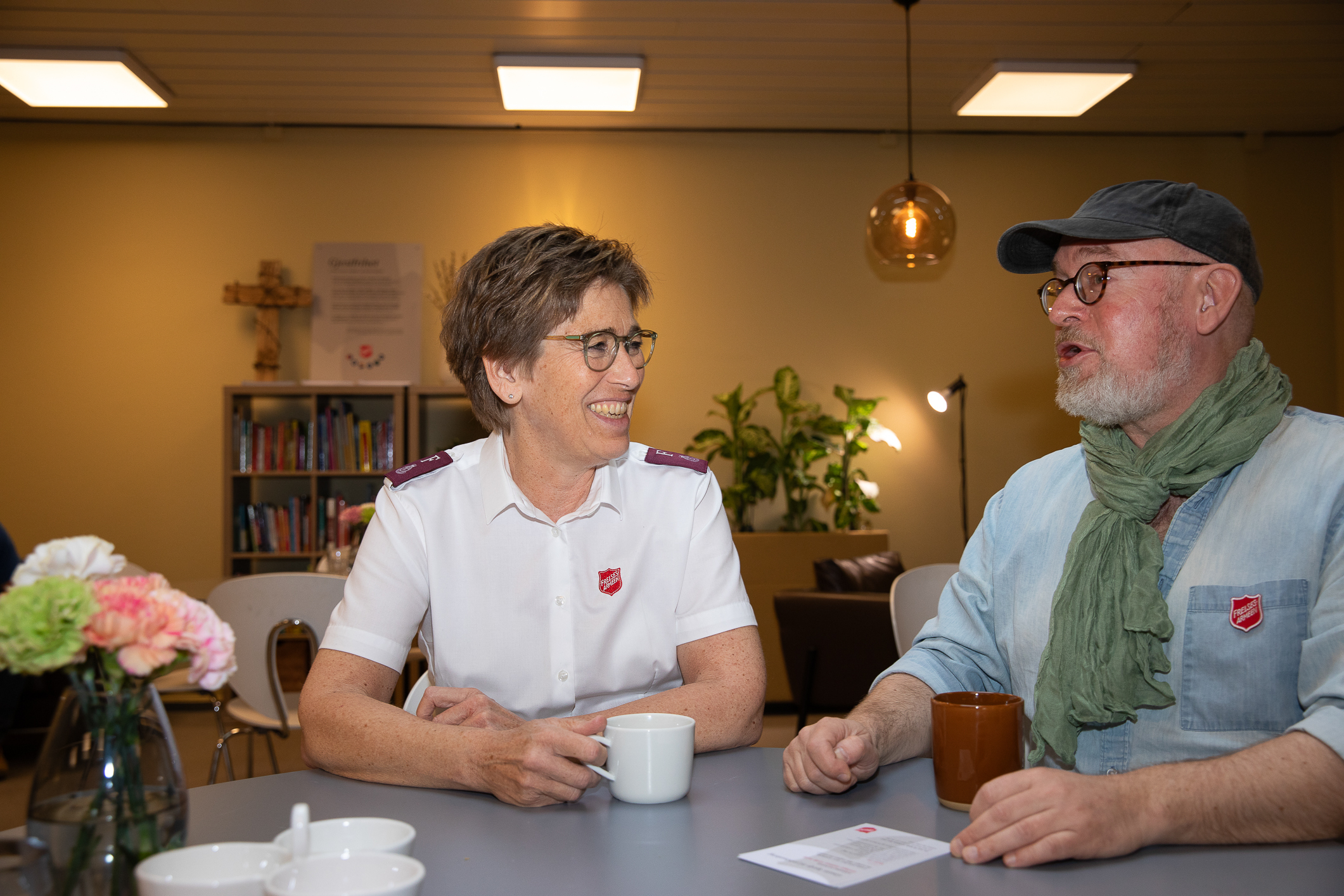 En middelaldrende kvinne med hvit uniformsskjorte, sitter ved et rundt bord og drikker kaffe med en mann med caps og lyseblå skjorte. Begge har Frelsesarmeens skjold på brystet.
