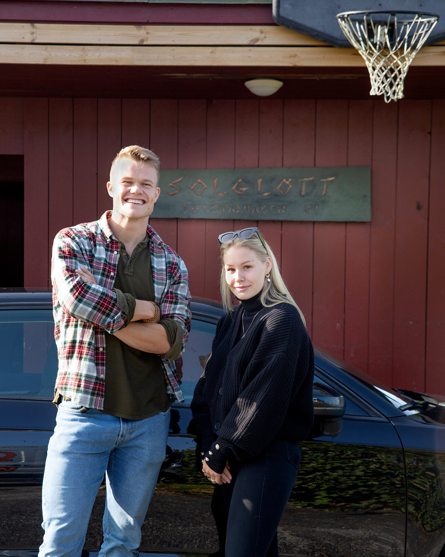 Bildet viser Endre og Siri som står foran en svart bil og en rød vegg med et skilt hvor det står Solgløtt. Begge smiler og ser i kamera.