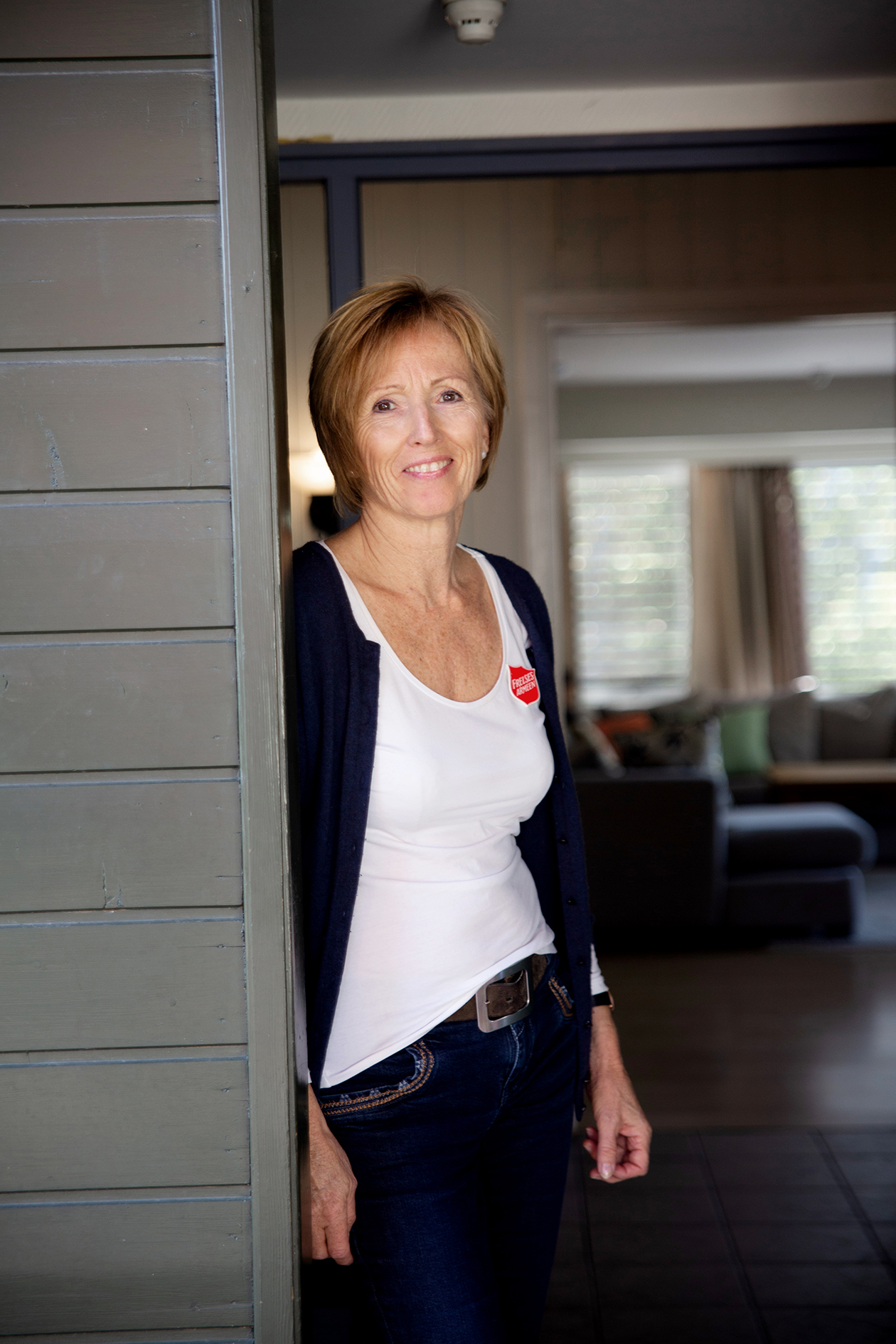 Bildet viser en kvinne med lyst kort hår som står i en døråpning. Hun har svarte bukser og hvit t-skjorte med Frelsesarme-skjold.