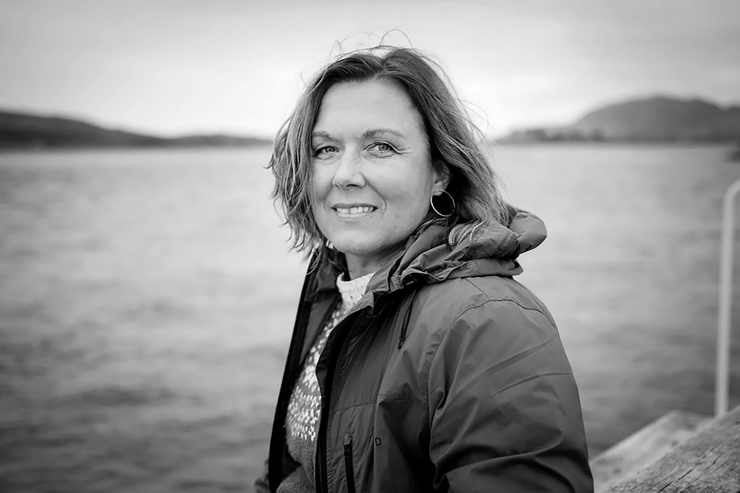 Bildet er et svart-hvitt nærbilde og viser en kvinne med halvlangt hår som står utendørs og ser mot kamera.