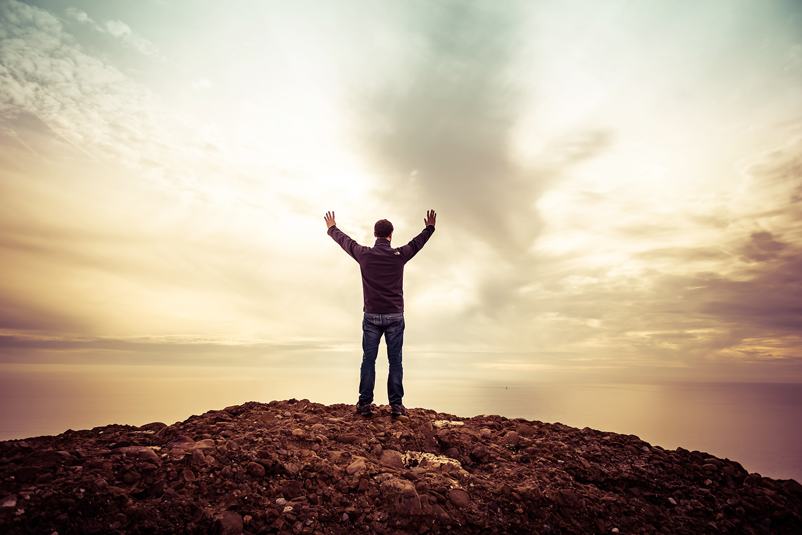 En mann står med ryggen til med hendene i løftet mot himmelen. Han står på en bakketopp i solnedgang