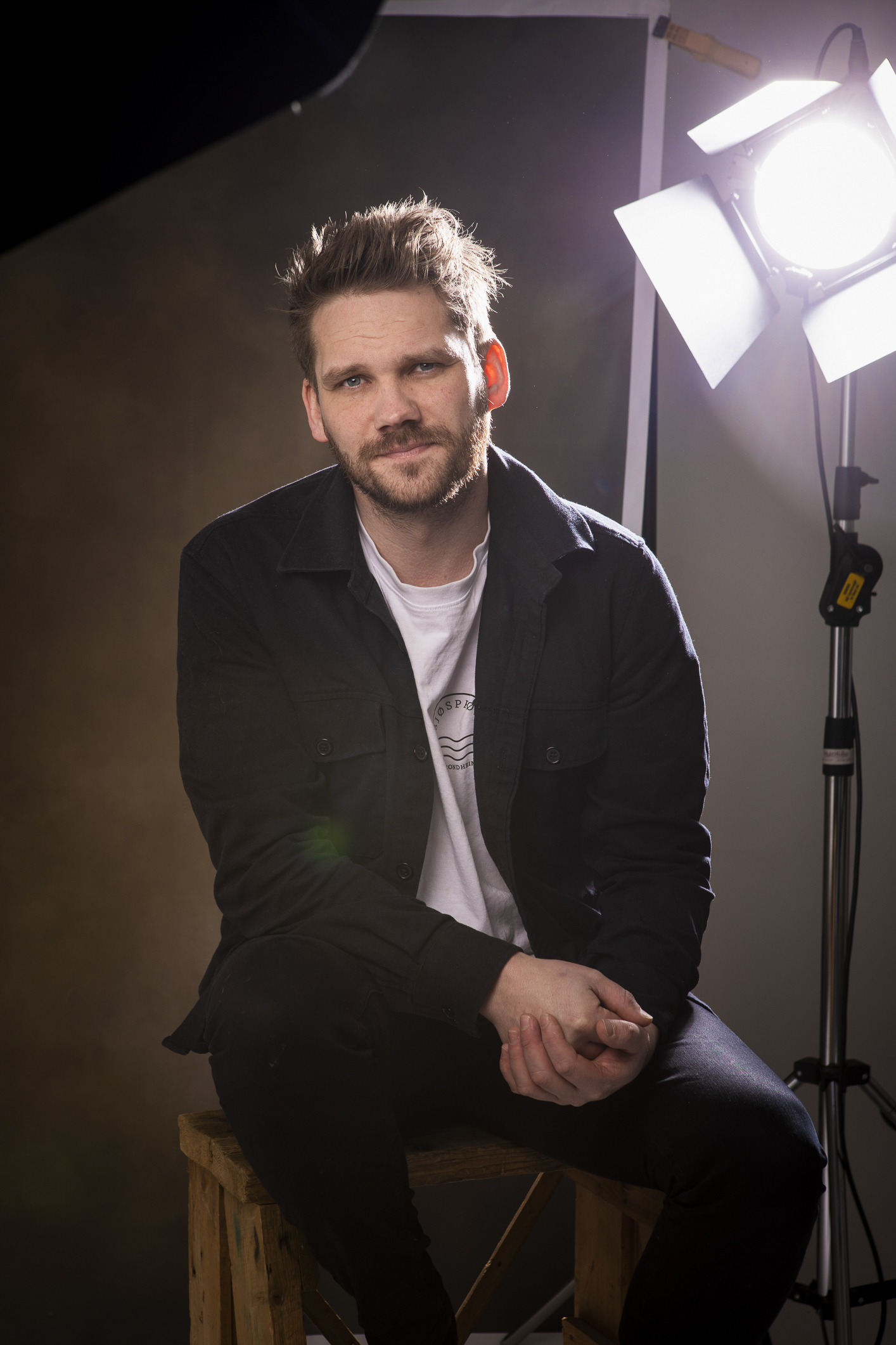 En ung mann med kort hår og skjegg sitter på en krakk i et brunt rom med hendene samlet foran seg. På høyre side står en foto-lampe og lyser.