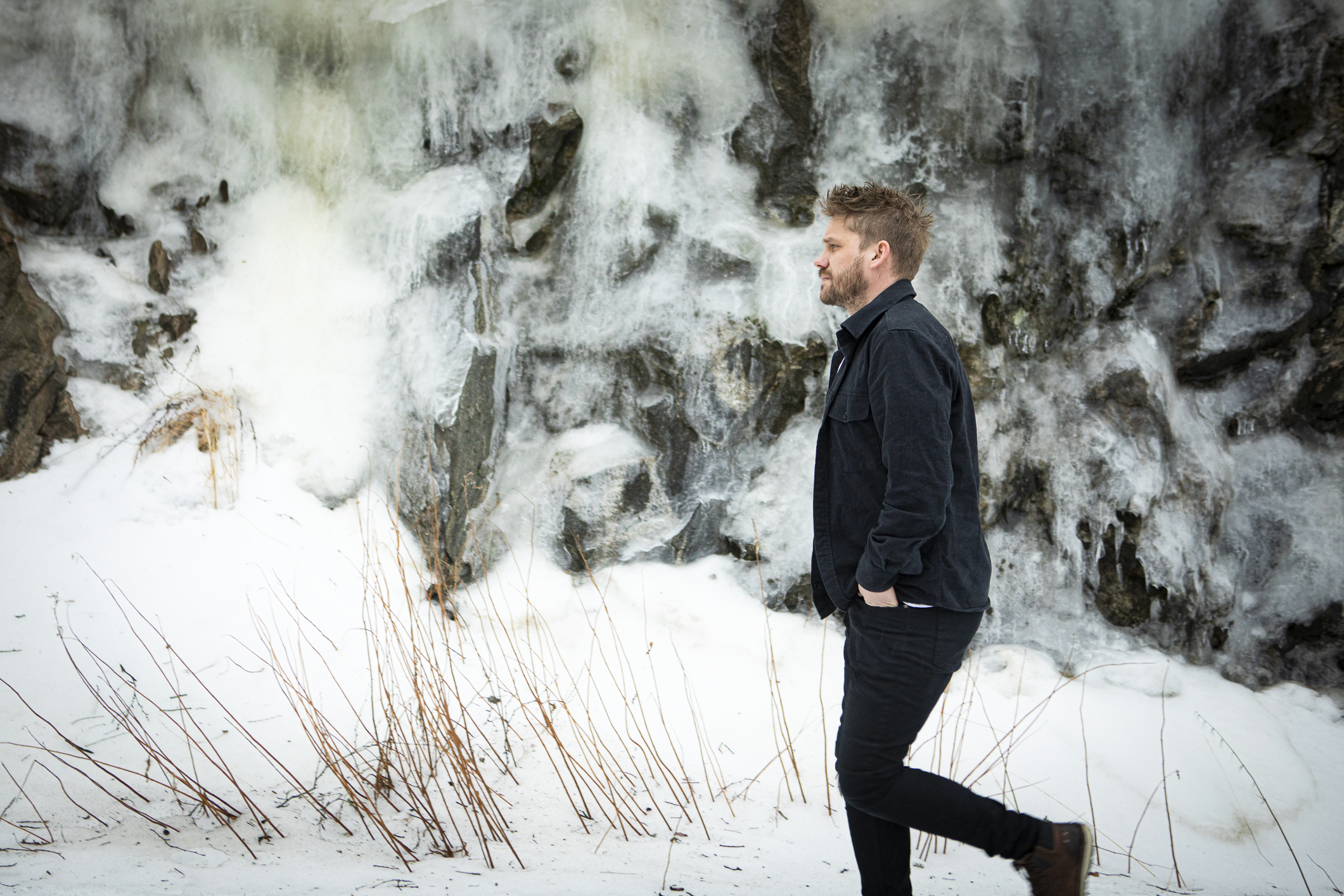 Hann med mørkt blondt hår og skjegg går ute med mørke klær og hendene i lomma.  Ute er det vinter og snøen henger tungt på trærne i bakgrunnen.