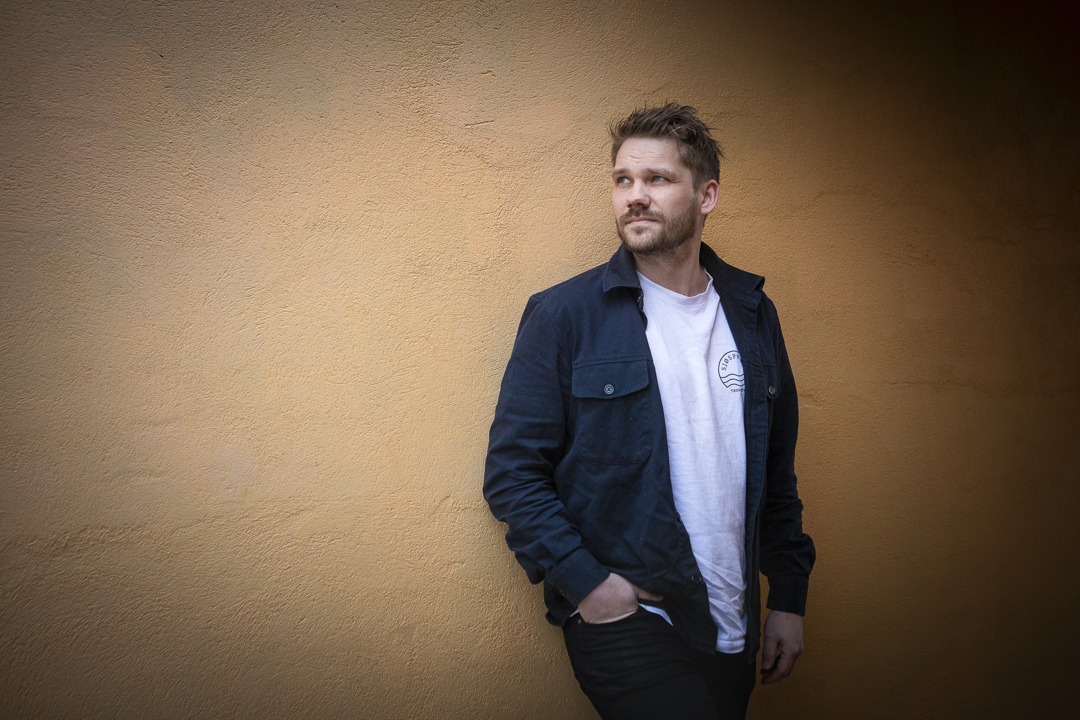 En ung mann med kort hår og skjegg står lent mor en gul vegg. Han har hvit T-skjorte inni en åpen marineblå, åpen jakke. Mannen har stylet hår og hendene i lommene.