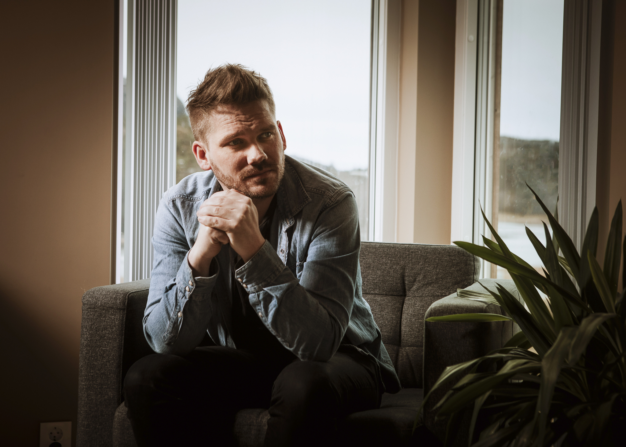En ung mann med kort hår og skjegg sitter i en brun sofa i et brunt rom med hendene samlet foran seg.