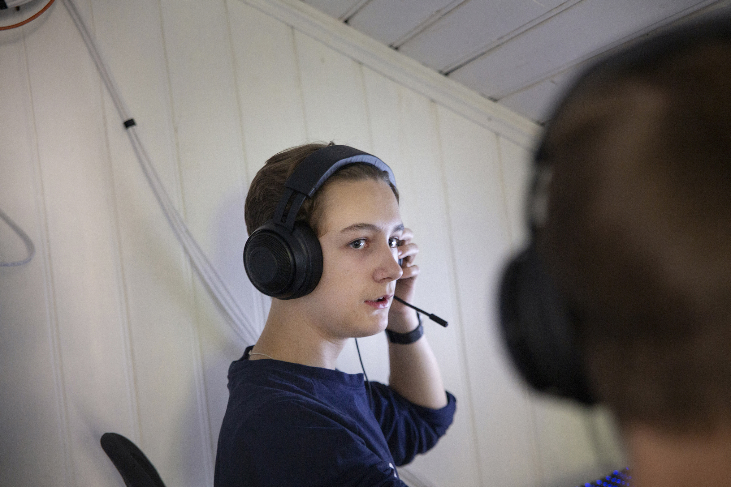 Det første bildet viser en ung mann som sitter og smiler mot en dataskjerm, han har flyttet headsettet bort fra ørene. Det andre bildet viser en annen ung mann som sitter med headsett og snakker med en annen person som kan skimtes i forgrunnen.