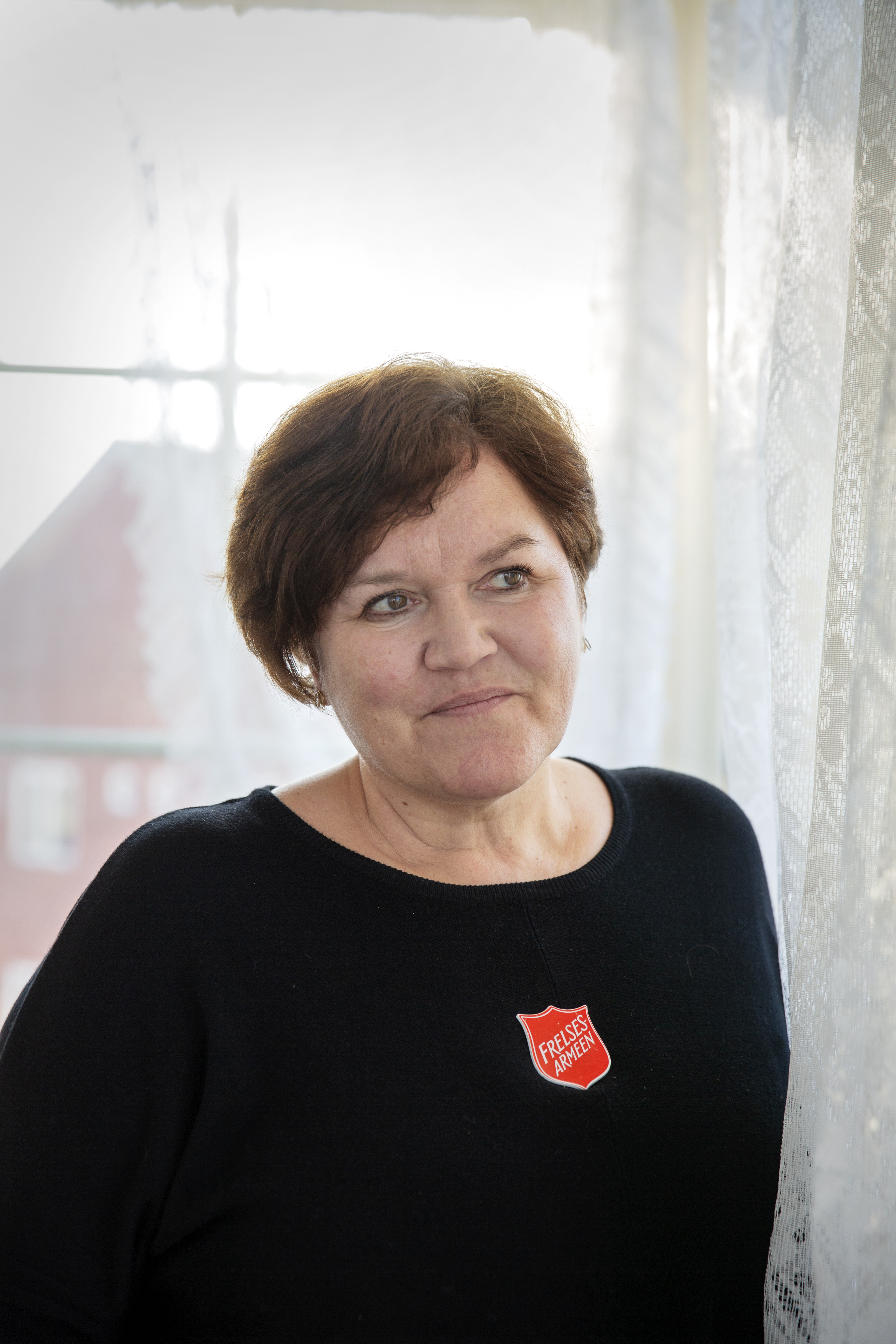 Til venstre ser vi Bernt Olaf Ørnes. Han har på seg Frelsesarmeens offisersuniform. Hen lener seg mot en dørkarm med armene i kors og smiler. Mona Rindal Rasmussen står foran et halvåpent vindu halvveis dekket av bondegardiner.Hun ser skrått forbi fotografen mot noe lenger borte.