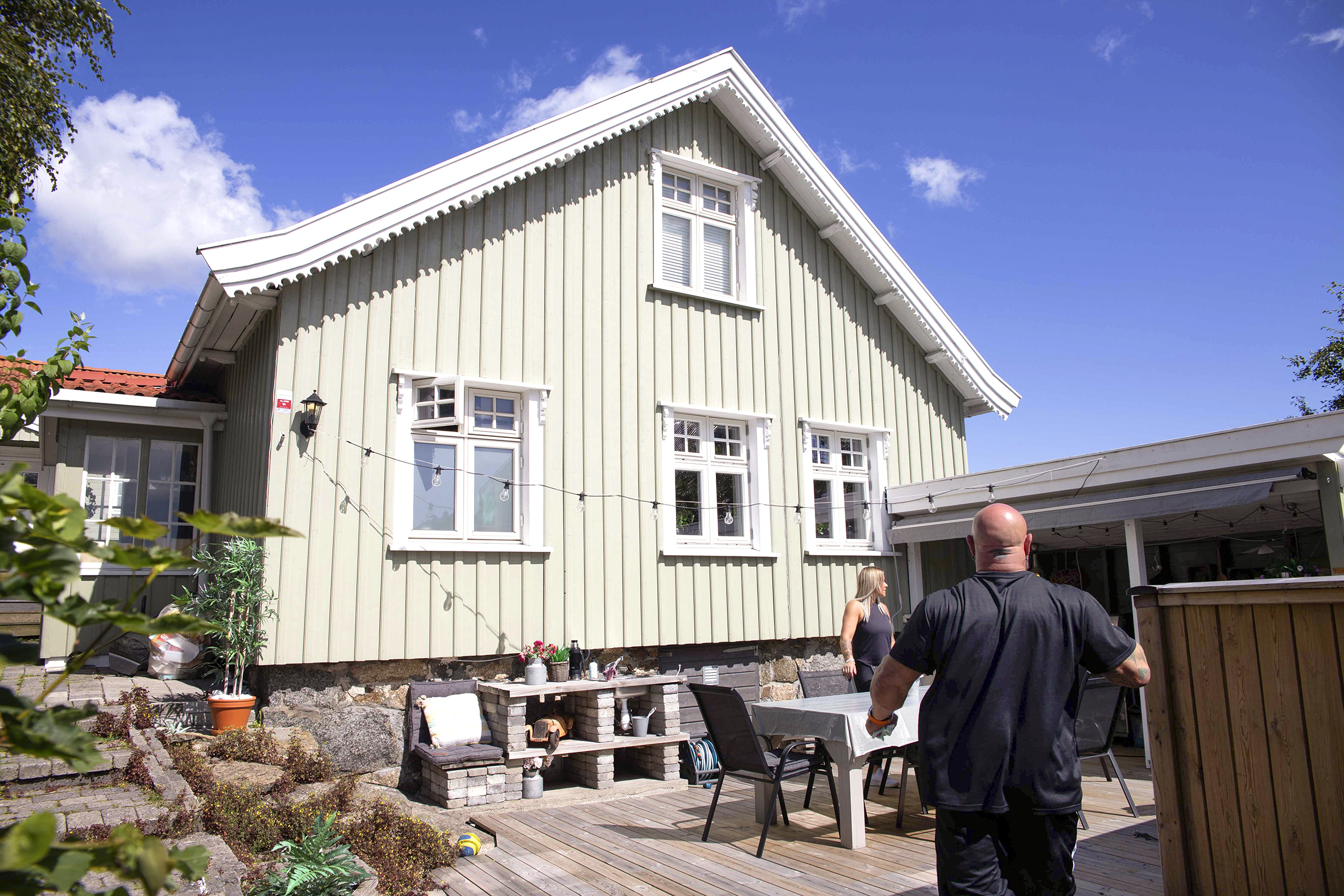 Bildet er tatt på avstand og viser en platting med bord og stoler utenfor et hvitt trehus. Thomas står nærmest med ryggen til og Ann-Hege sees i bakgrunnen.