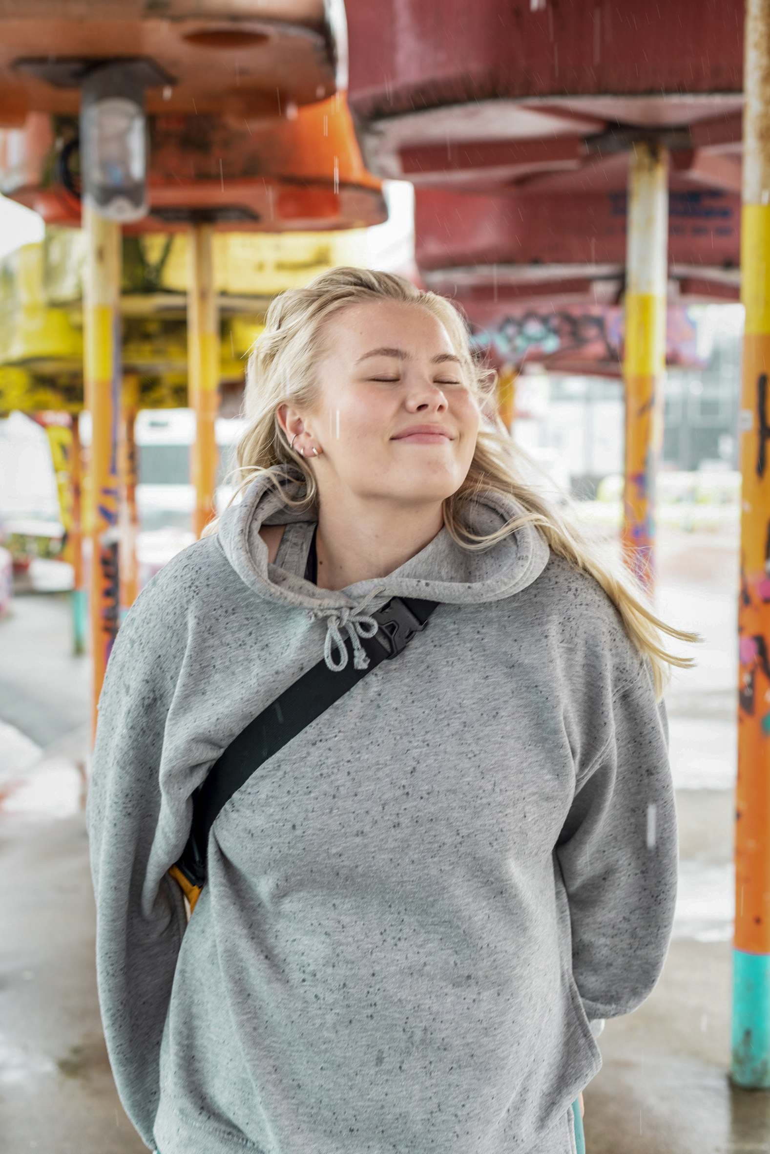 Bildet viser en ung kvinne som står med ansiktet vendt oppover, og smiler med lukkede øyne. Hun har lyst langt hår og grå hettegenser.