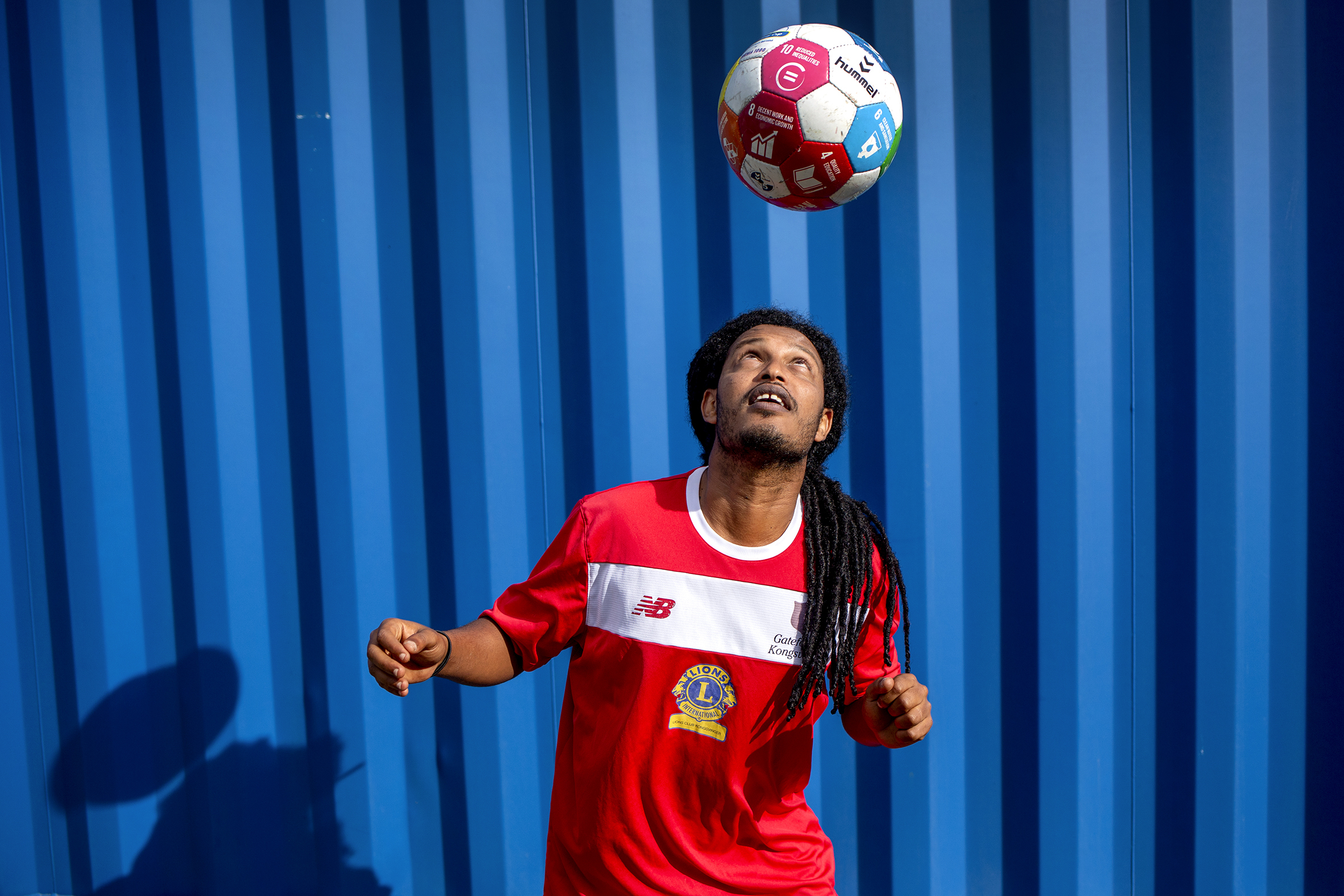 Bildet viser Atie som står foran en blå vegg. Han er klar til å heade ballen som er i lufta rett over ham. Han har rød fotballt-skjorte og langt hår i rastafletter.