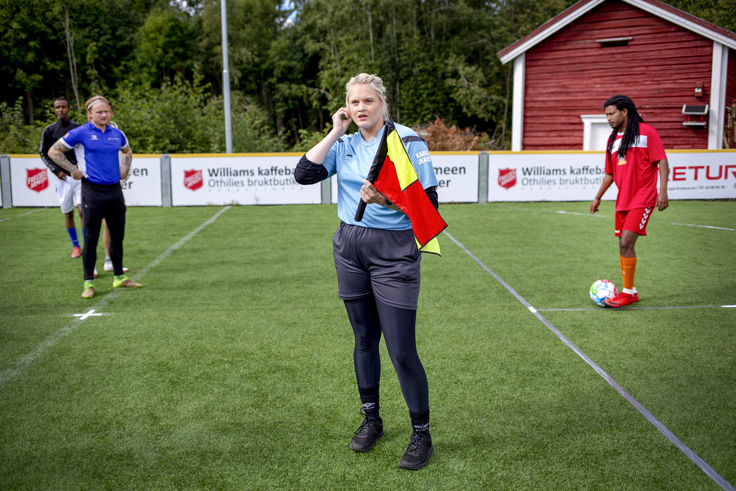 Bildet viser en ung kvinne med lyst hår, blå dommer-tskjorte og svart shorts og tights under. Hun står på banen og holder et rødt flagg.
