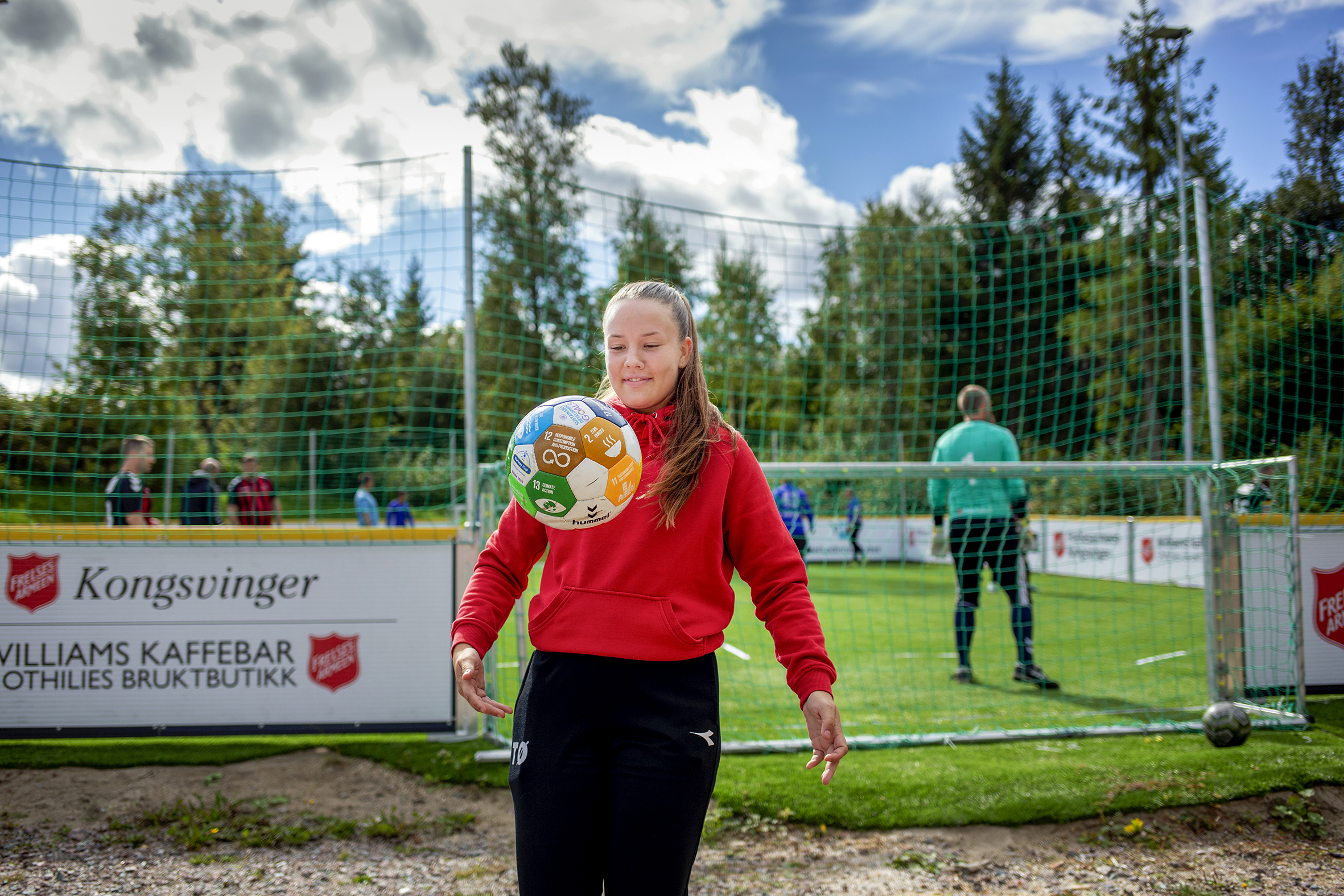Bildet viser en ung kvinne i rød genser og med lyst langt hår, som står og trikser med en ball, som er i lufta rett foran henne.