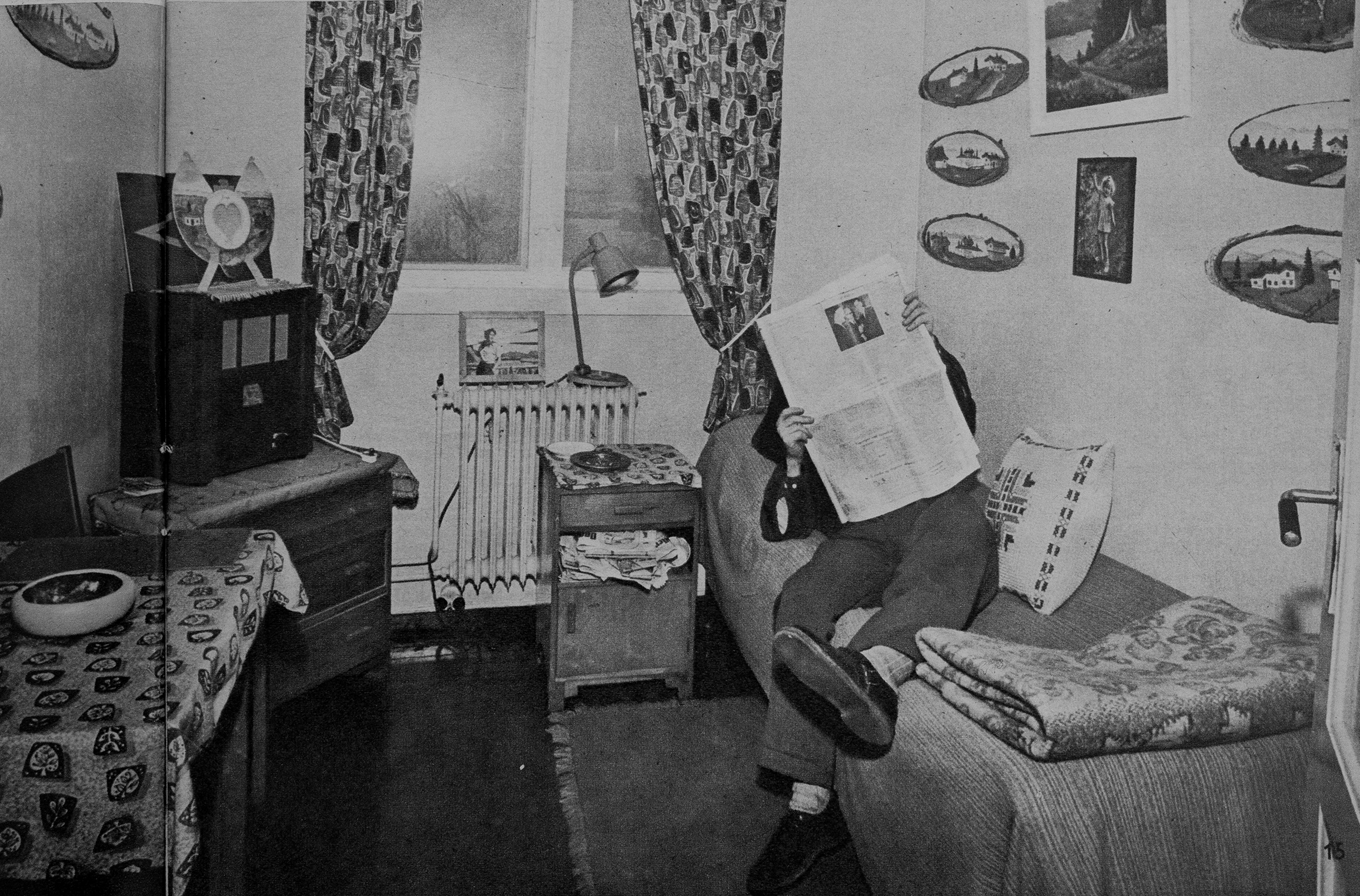 Et gammelt svart-hvitt bilde av en mann som ligger på en seng på et lite rom, ansiktet hans er skjult bak avisen han leser. Han har mange bilder på veggene og forskjellig pynt og andre gjenstander på rommet.