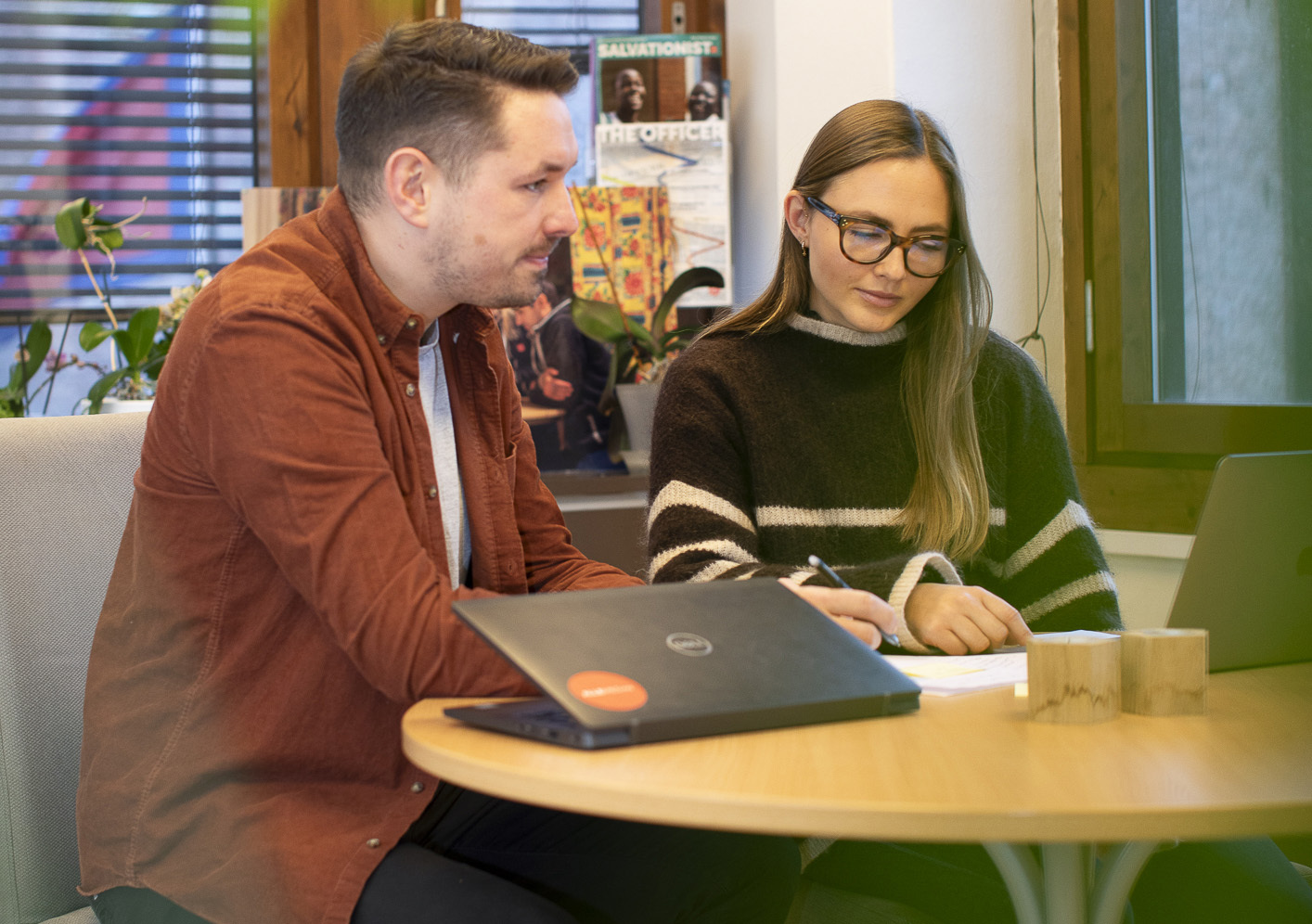 En ung mann og en ung kvinne sitter på et rundt bord og jobber på hver sin pc De sitter på et kontor med vinduer og en plante i bakgrunnen