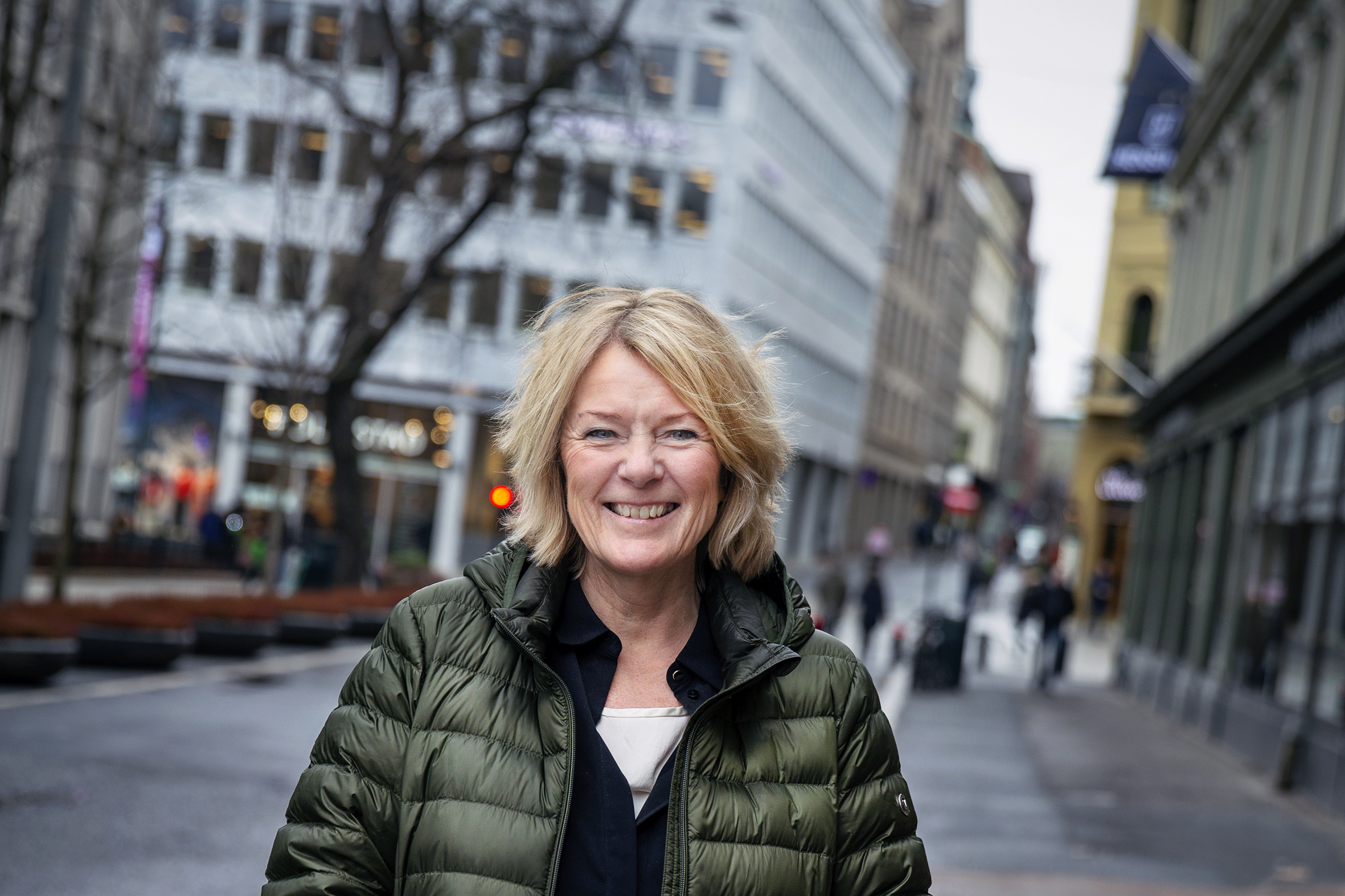 Kristin Clemet står i en av Oslo gater. Hun i fokus nærmest kamera, og smiler bredt. Hun har på seg en tynn boblejakke med grønnskjær.