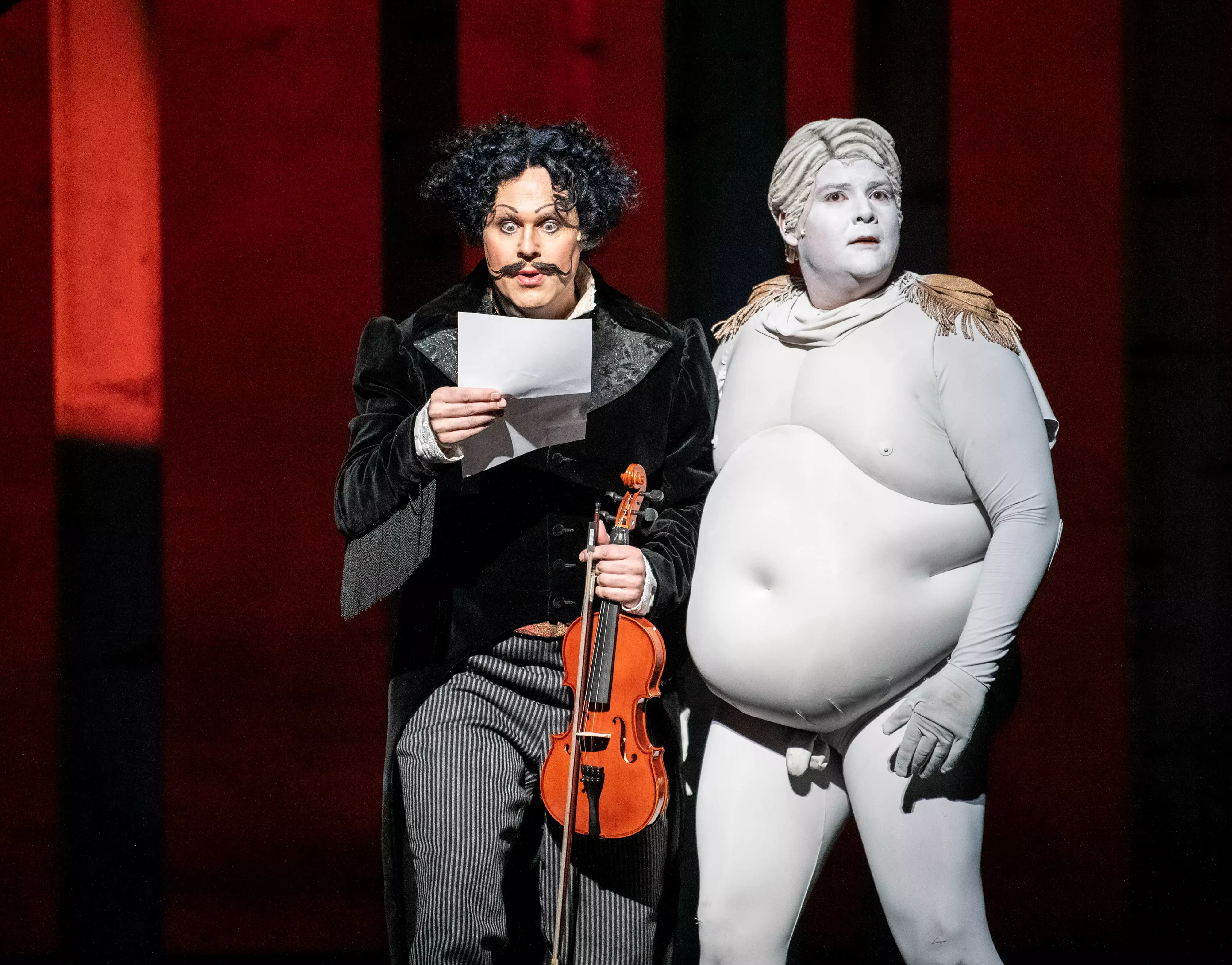 To menn står sammen på scenen. Den ene, iført svart dress, holder en fiolin og den andre står i en hvitmalt "fatsuit".