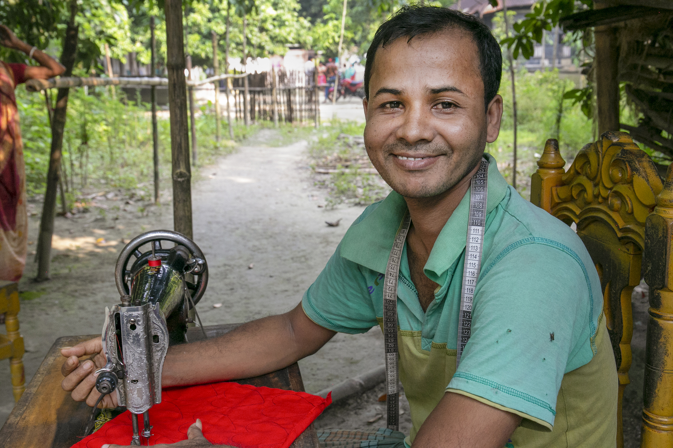 Bildet viser en mann fra Bangladesh som sitter ved en symaskin. Bildet er tatt fra siden, han snur seg og smiler mot kamera. I bakgrunnen sees trær og gress.