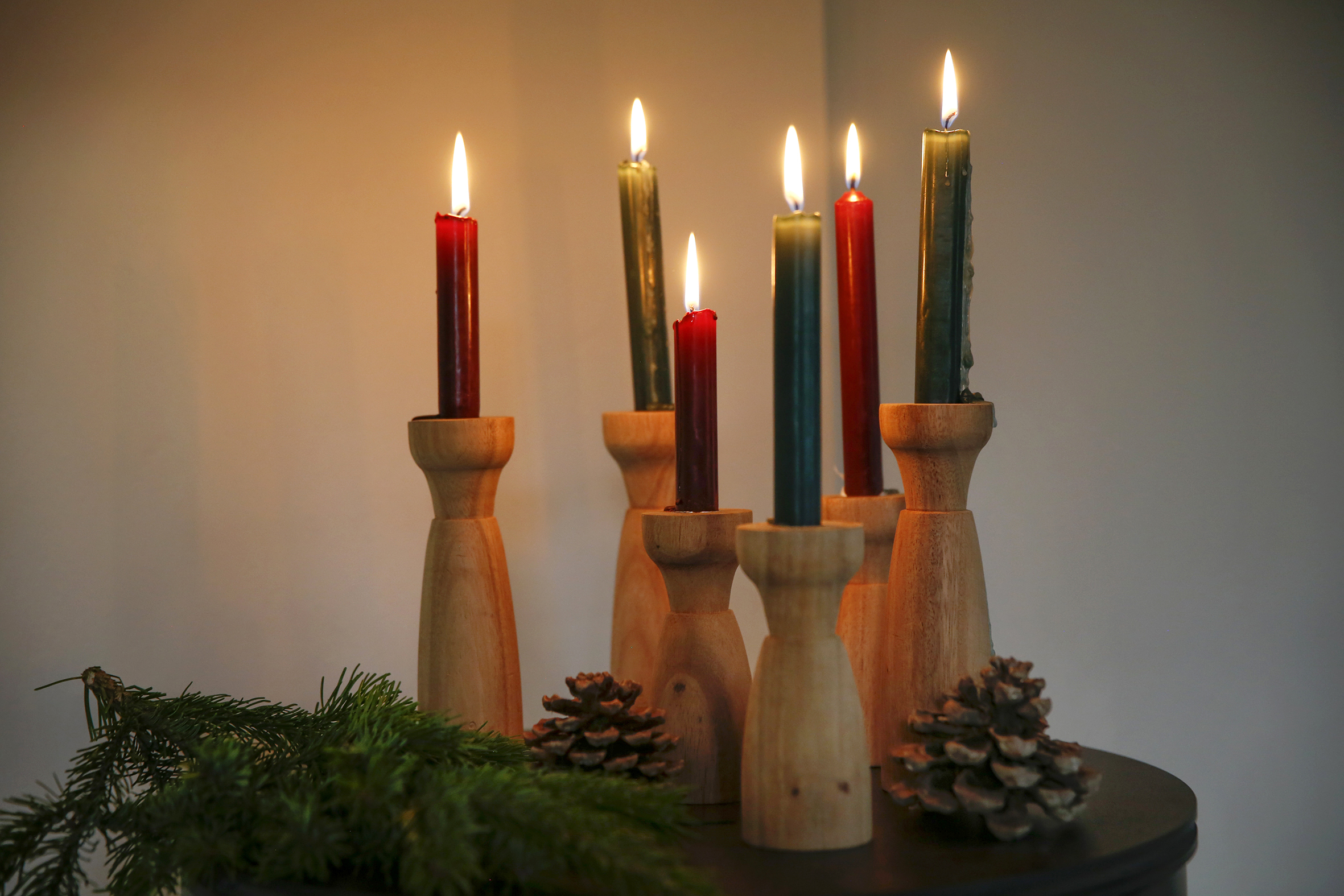 Bildet viser seks lysestaker i tre, med tente stearinlys i og granbar og kongler rundt.