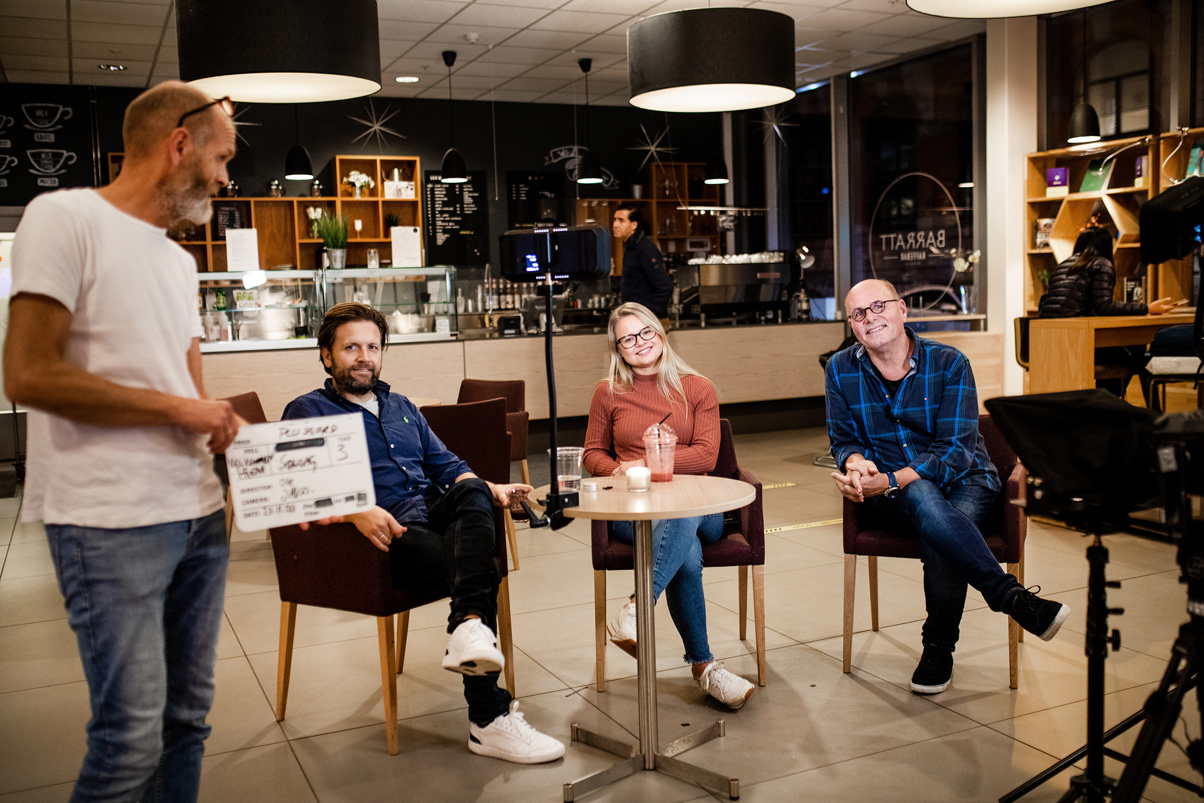 Tre personer sitter rundt et bord og smiler mot kamera mens produsenten står i forgrunnen, klar til å starte opptak