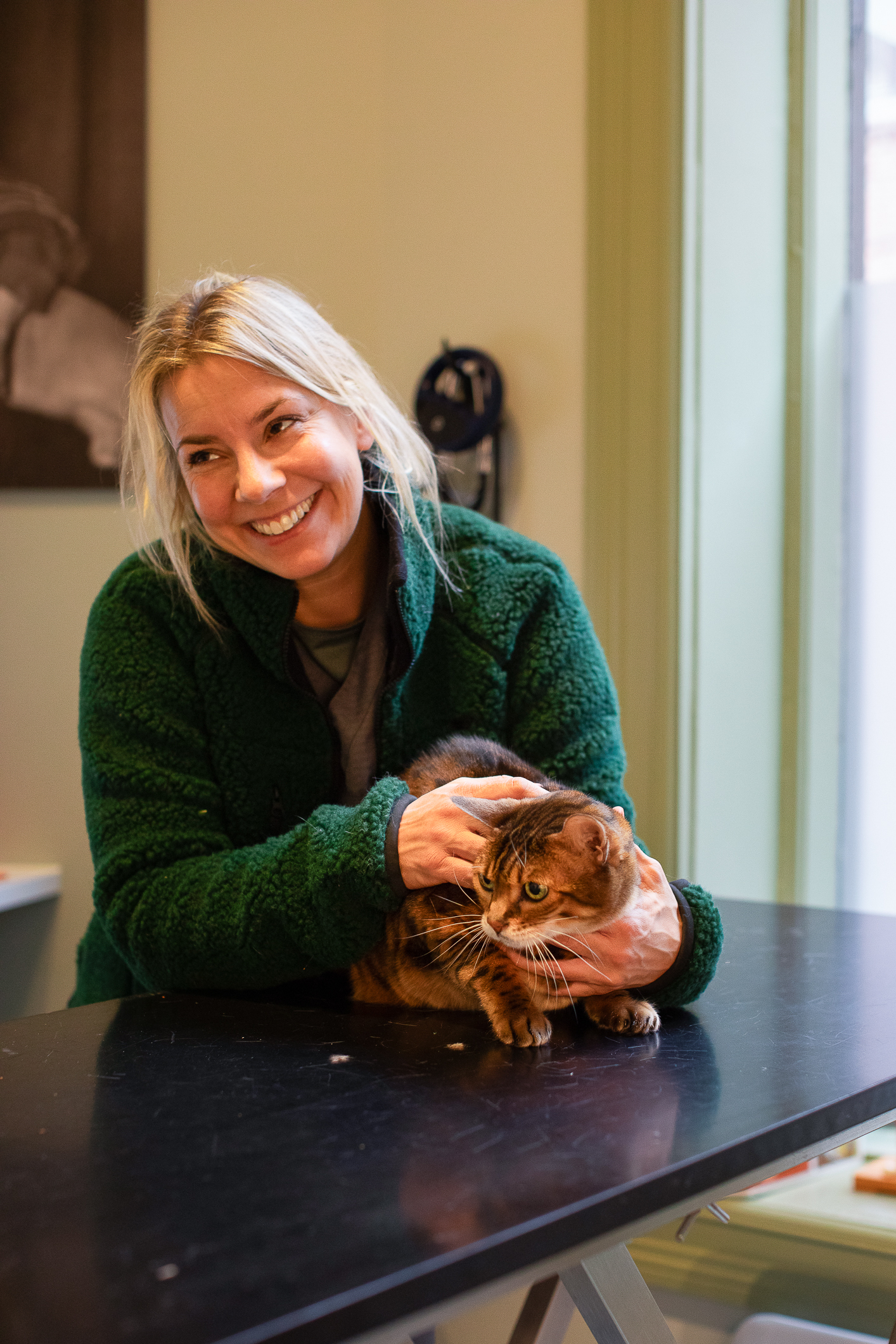 Kvinne med grønn fleecegenser klapper en bengalkatt oppå et bord med sort overflate hos veterinæren.