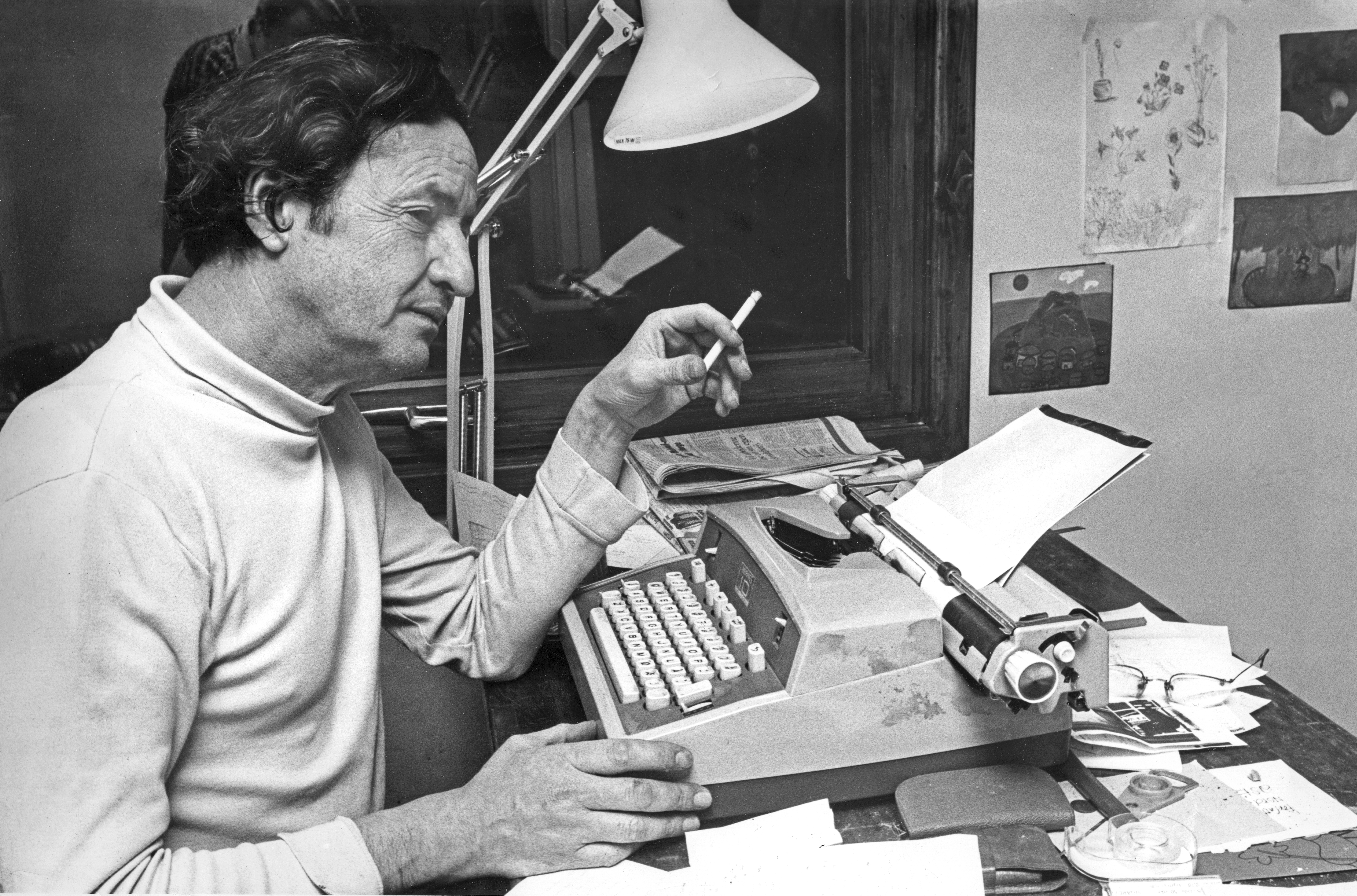 Jens Bjørneboe sitter ved sin skrivemaskin. Han holder en sigarett i hånden.