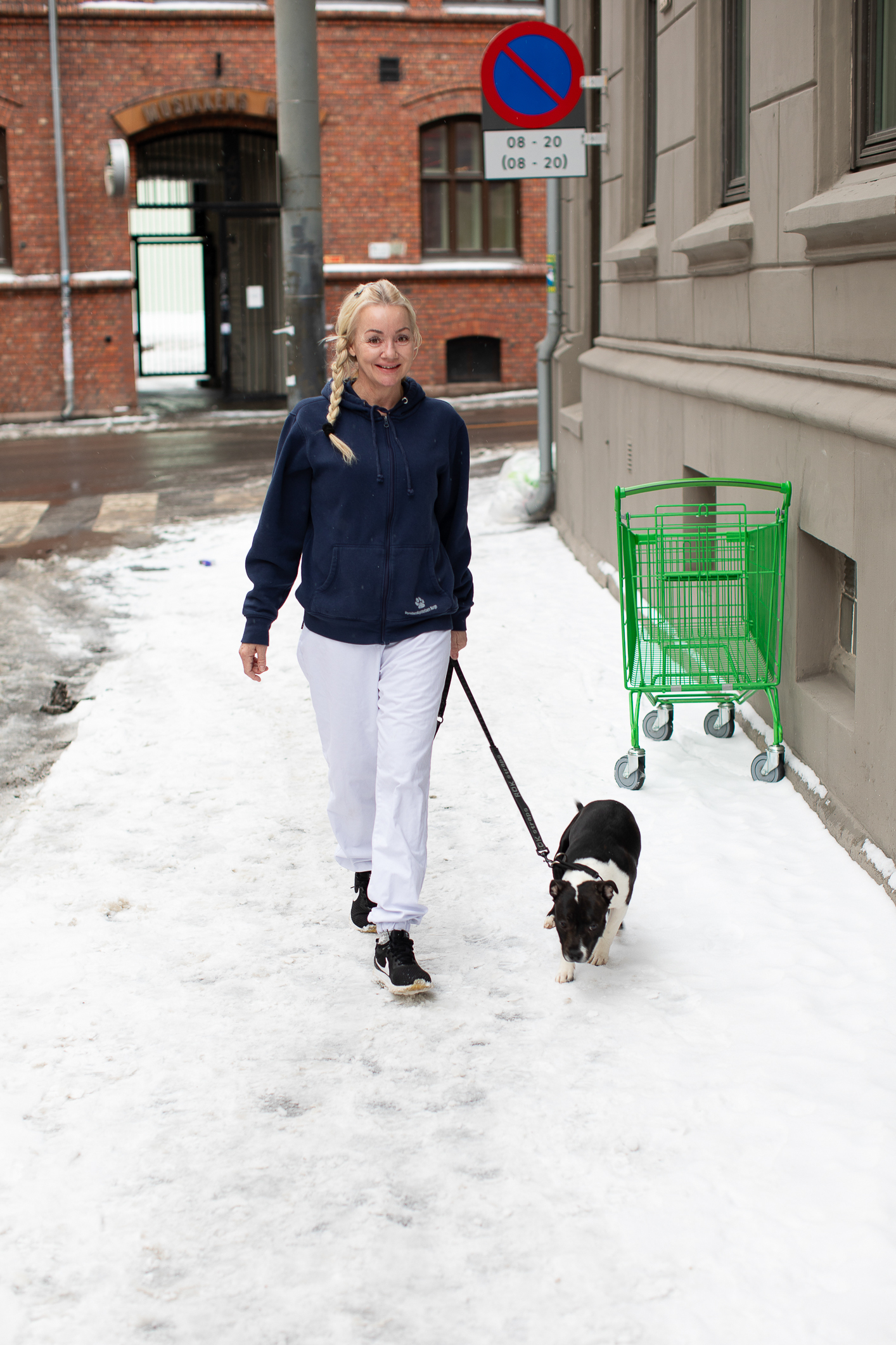 Ung kvinne med lys flette, blå fleece-genser og hvit bukse, går rundt kvartalet og lufter en liten sort og hvit hund. Hun her joggesko. Ute på gaten er det snø på fortauet.