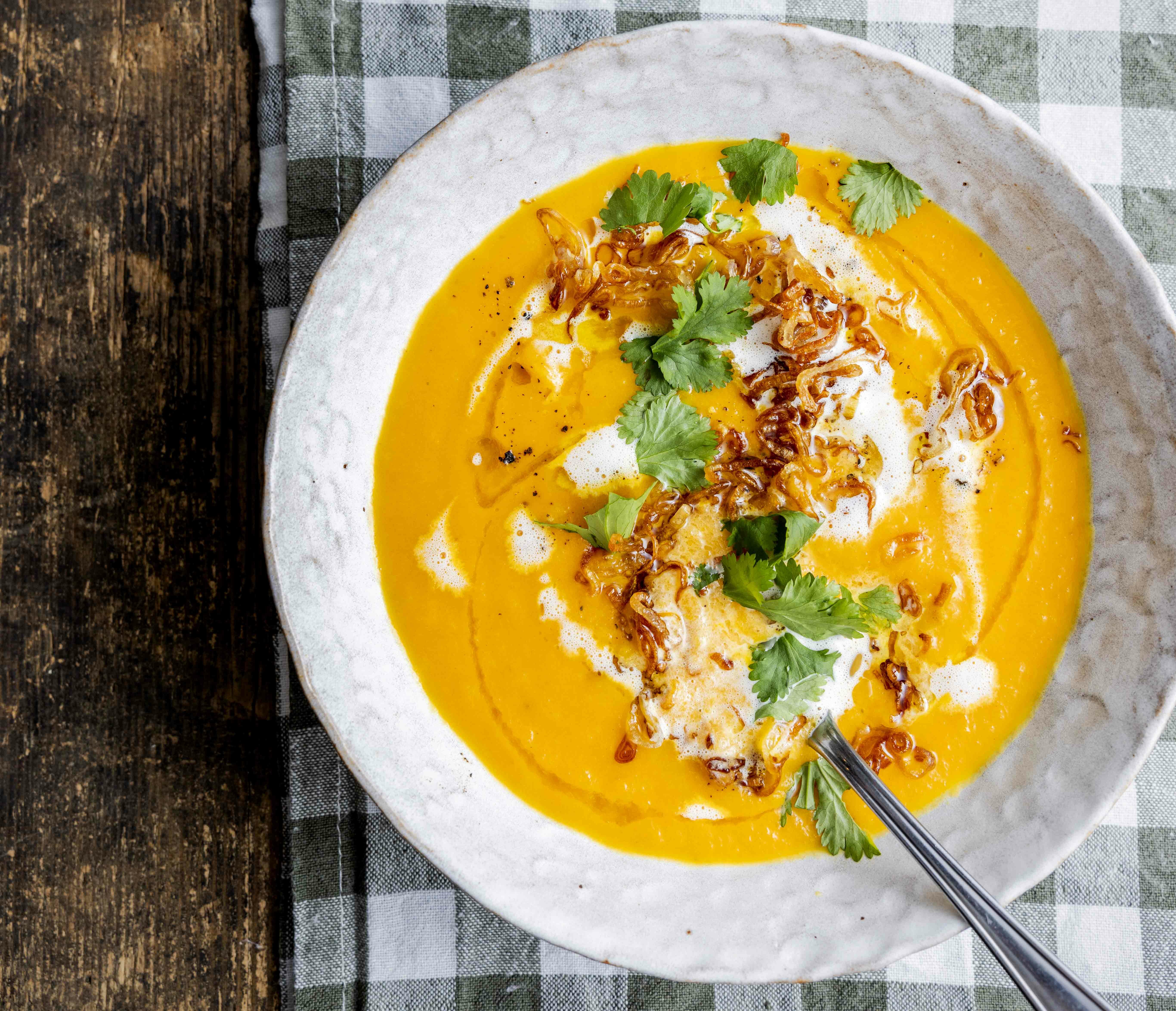 Foto: Nærbilde av en hvit suppebolle med oransje gulrotsuppe i. Bollen står på en rutete duk. Suppen er toppet med persilleblader, hvit kokosmelk og stekt løk.