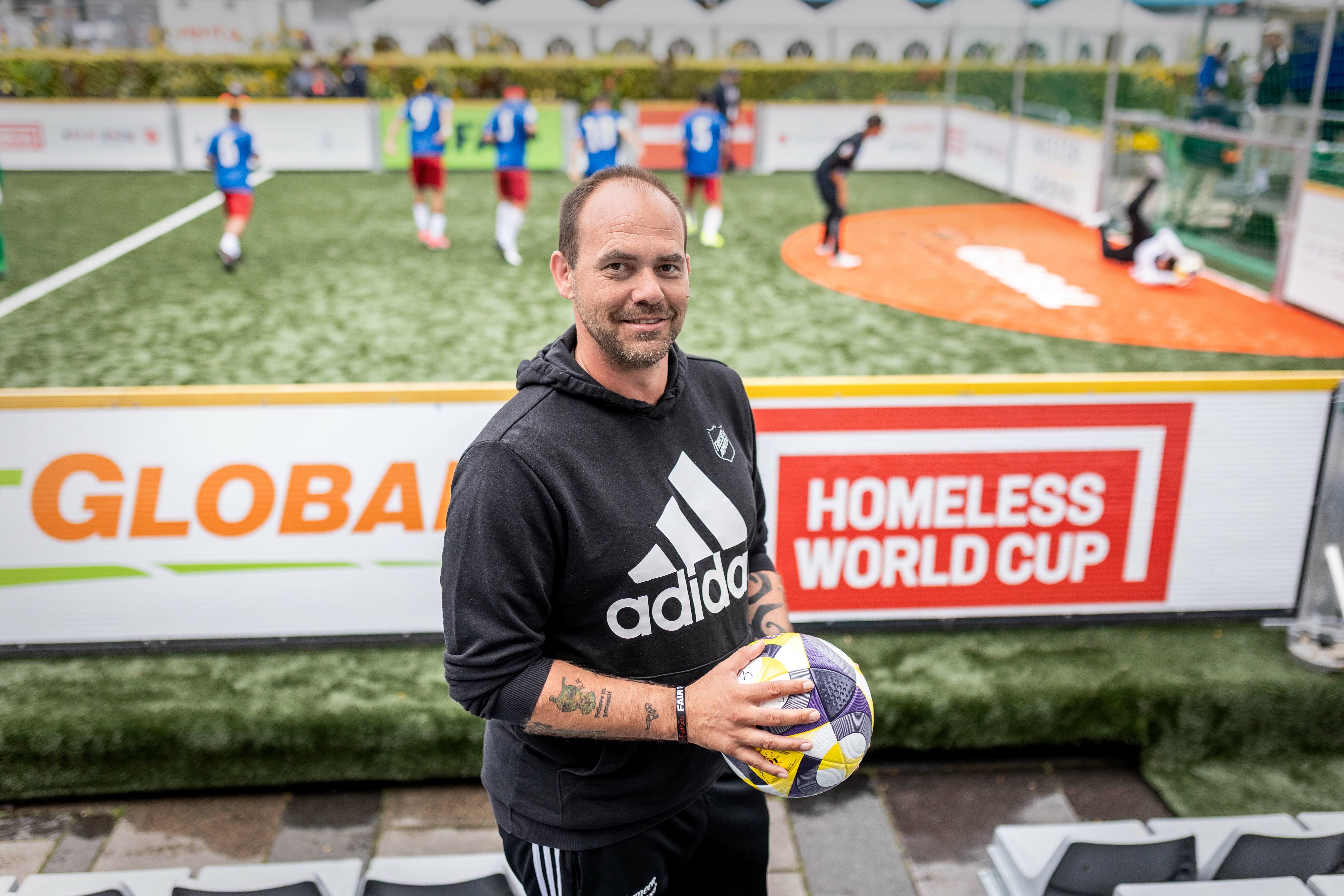 Foto: En mann i svart Adidas-genser og står litt oppe i tribunen under VM, han smiler mot kamera og holder en ball. Bak ham sees banen med spillere i blå t-skjorter og røde shorts som spiller en kamp.