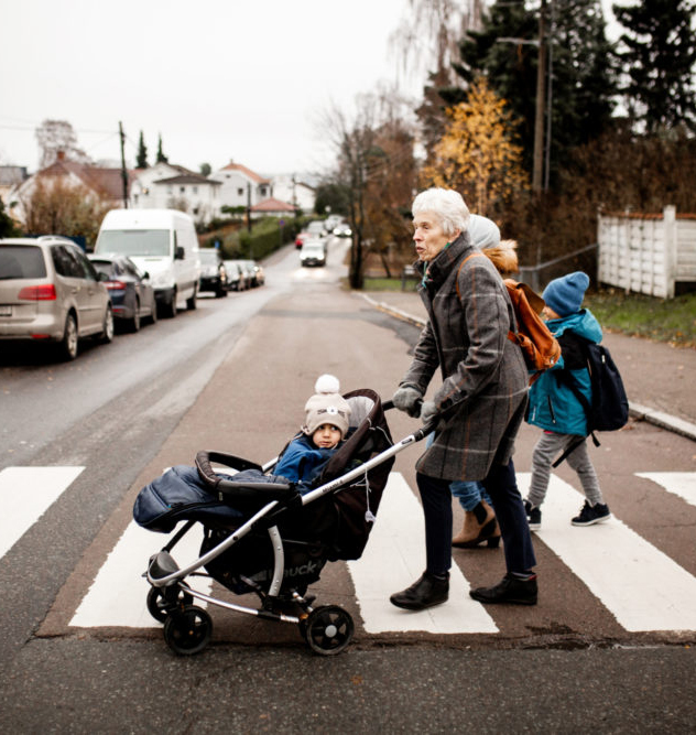 Eldre kvinne kjører barnevogn med liten gutt i. Ved siden av går guttens mamma og bror.