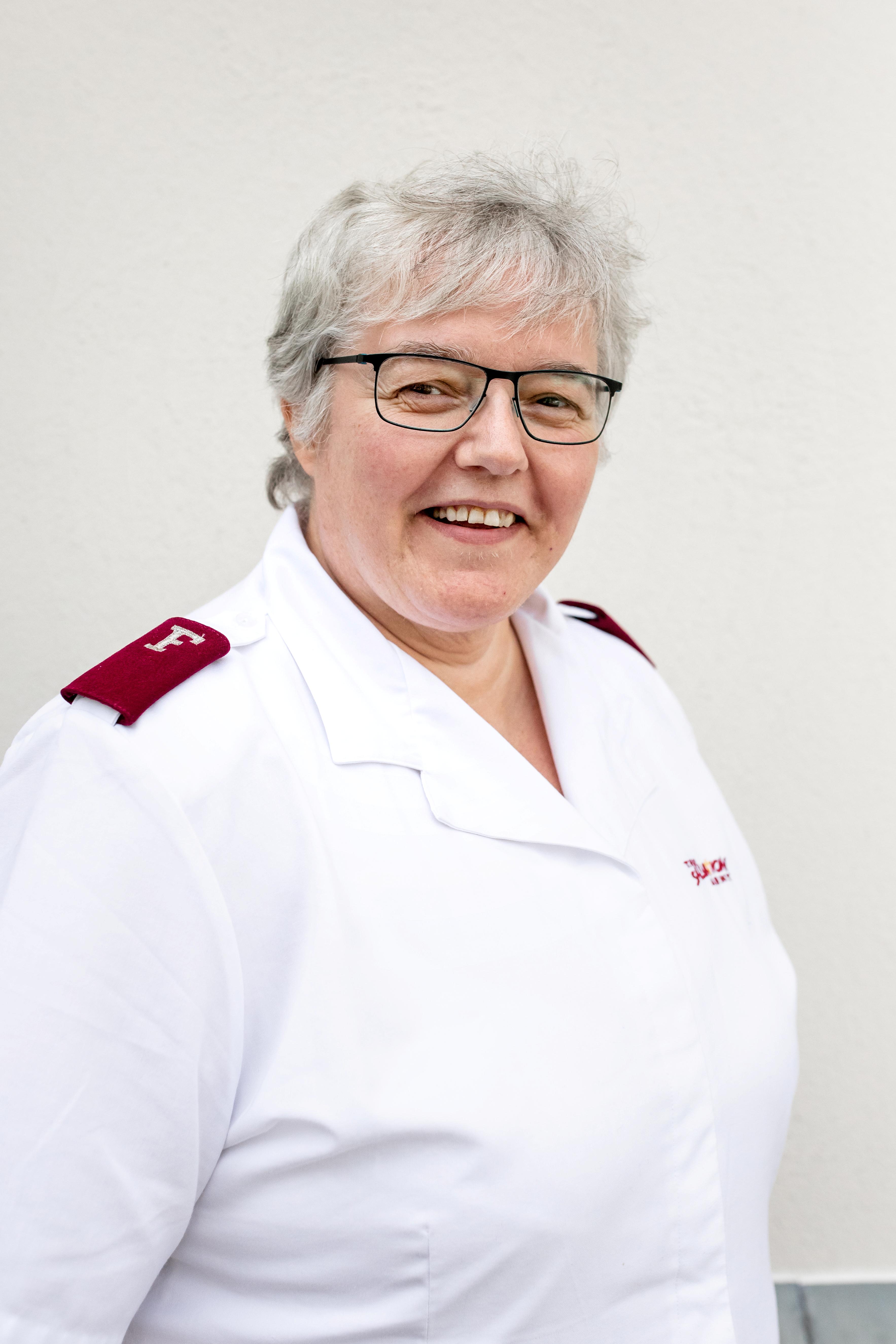 Portrett av offiser i Frelsesarmeen Marianne Spor. Her er hun iført hvir uniformsskjorte med lilla offisersklaffer på skuldrene. Hun har brille, grått, kort hår og ser i kameraet med et smil