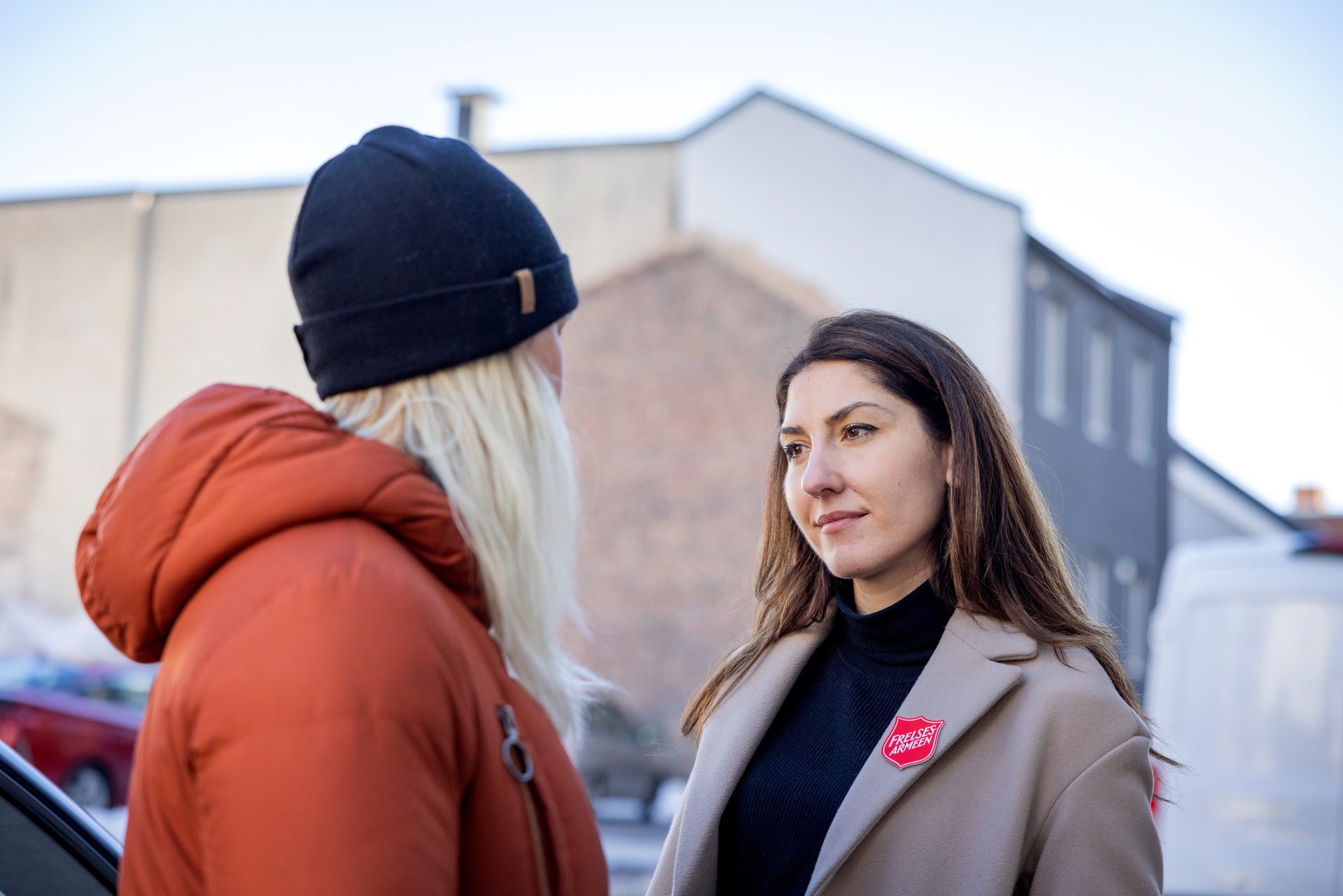 Foto: Kvinne med mørkt, langt hår og logo fra Frelsesarmeen på kåpeslaget, står og snakker med en ung, blond dame med lue og boblejakke på.