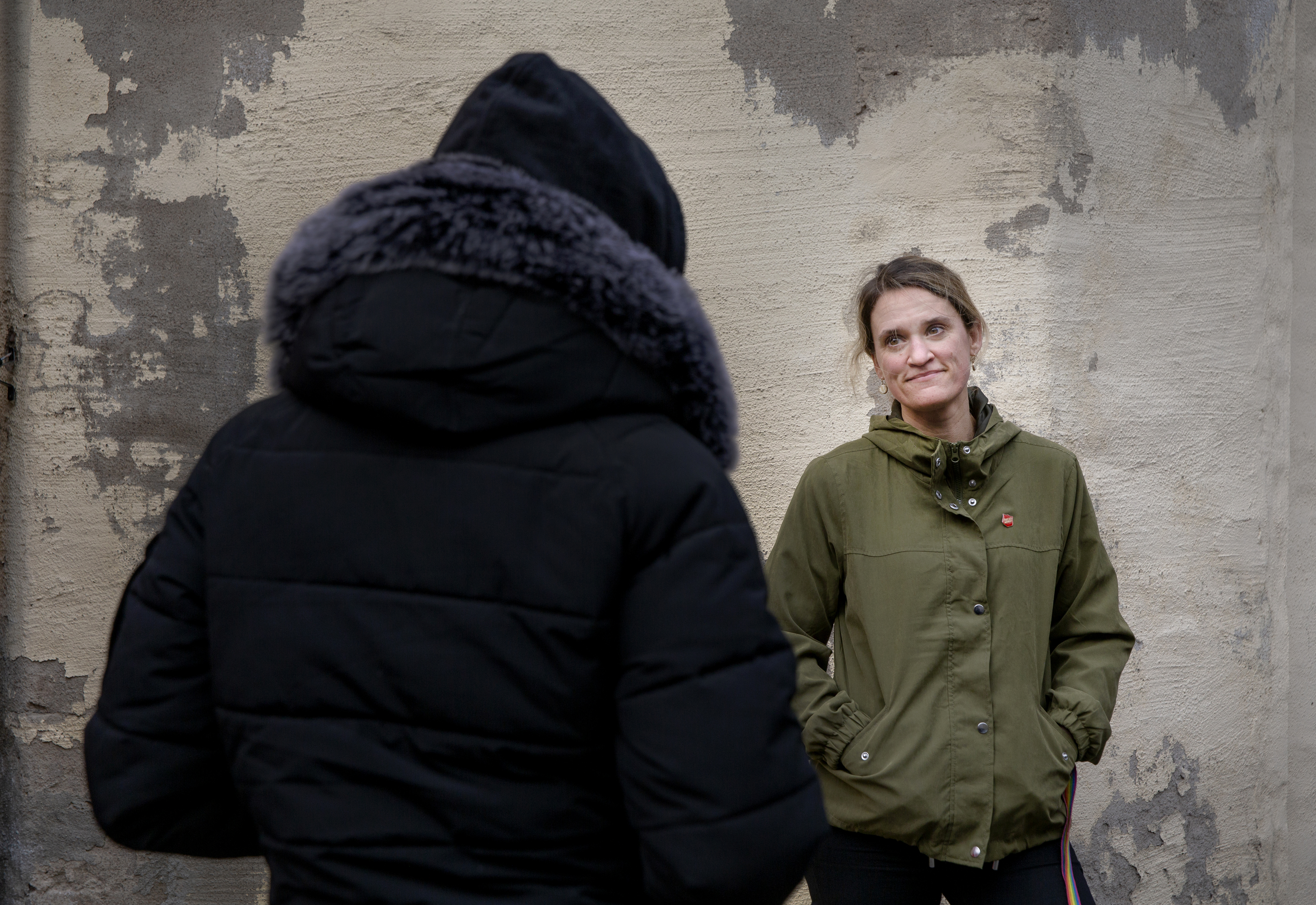 To voksne som prater sammen. Den ene kvinnen har på seg grønn ytterjakke med en grå murvegg i bakgrunn. Den andre har på seg sort ytterjakke, med ryggen til kamera.