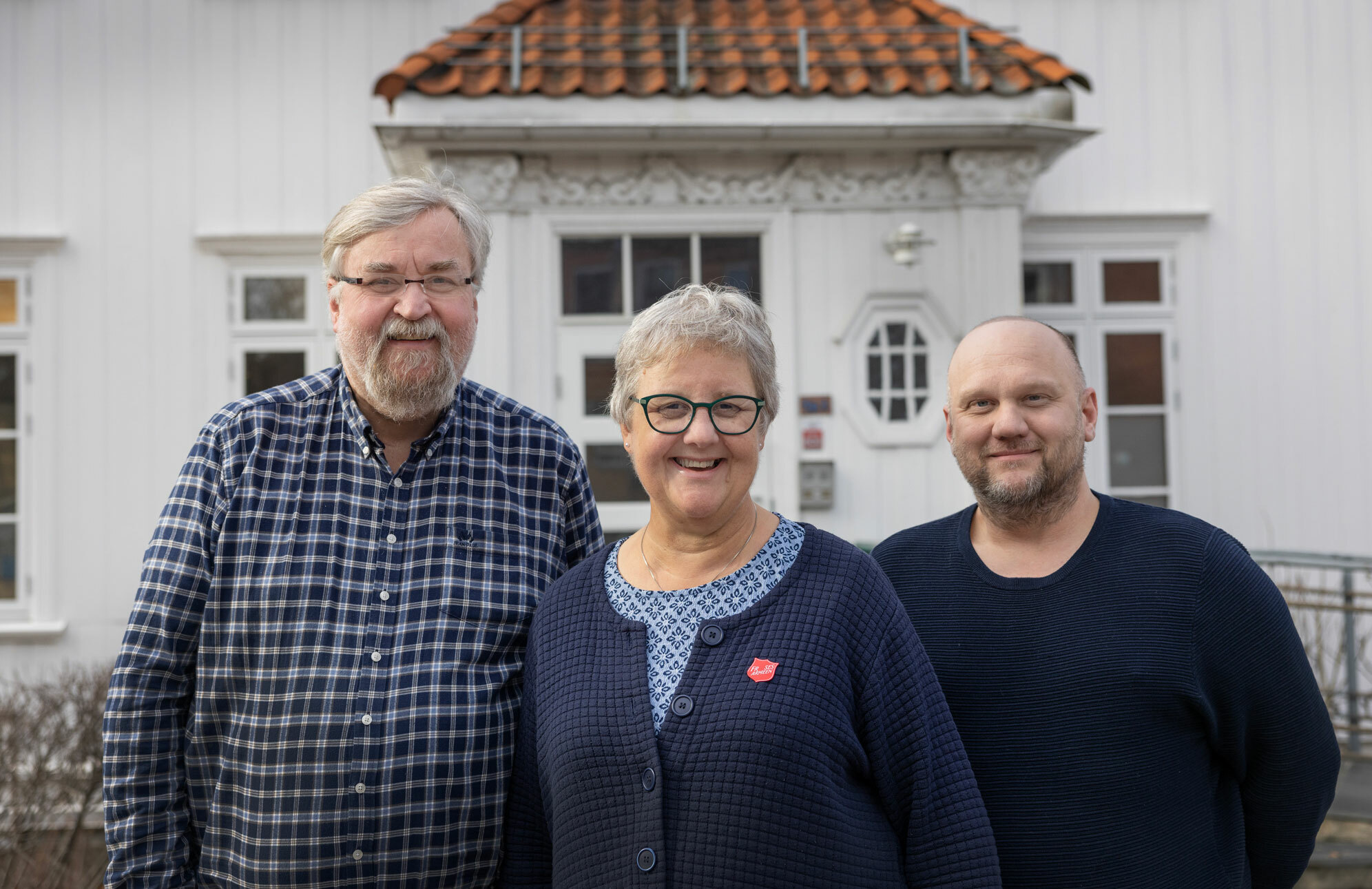 Foto: To menn med en kvinne mellom seg står oppstilt for fotografen utenfor et gammelt, hvitt trehus. Alle er i 50-60-årene.