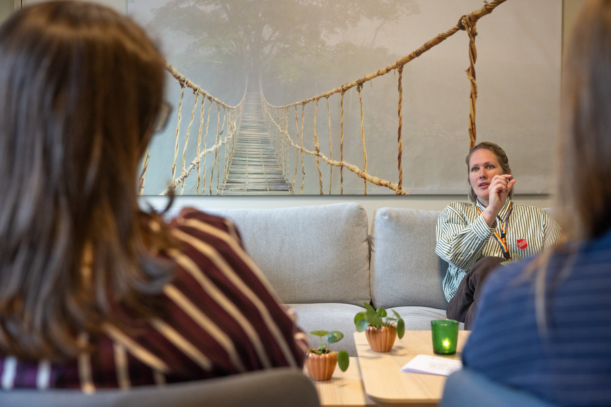 Foto: En kvinne med lyst hår og grønn og hvit stripete skjorte sitter i en sofa og snakker med to kvinner. Bak henne er et stort bilde av en taubro. Bare bakhodene til kvinnene synes uskarpt i forgrunnen.