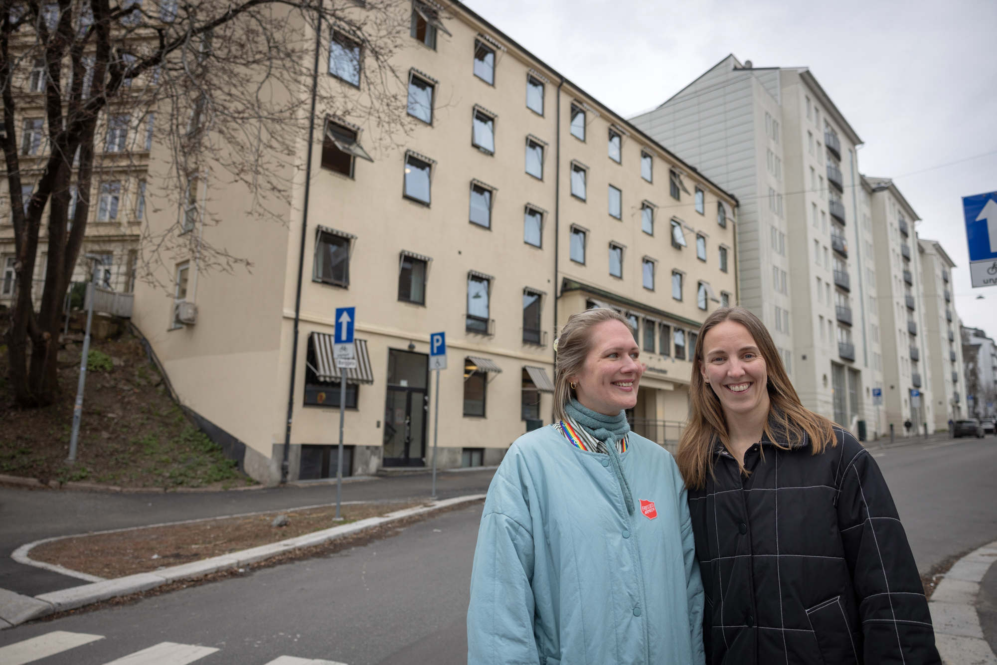 Foto: To kvinner står ute på gata. Den ene har lyst hår og lyseblå kåpe, den andre har mørkt hår og mørk, rutete jakke. Bak dem sees et stort lysegult murbygg og flere like, hvite blokker.