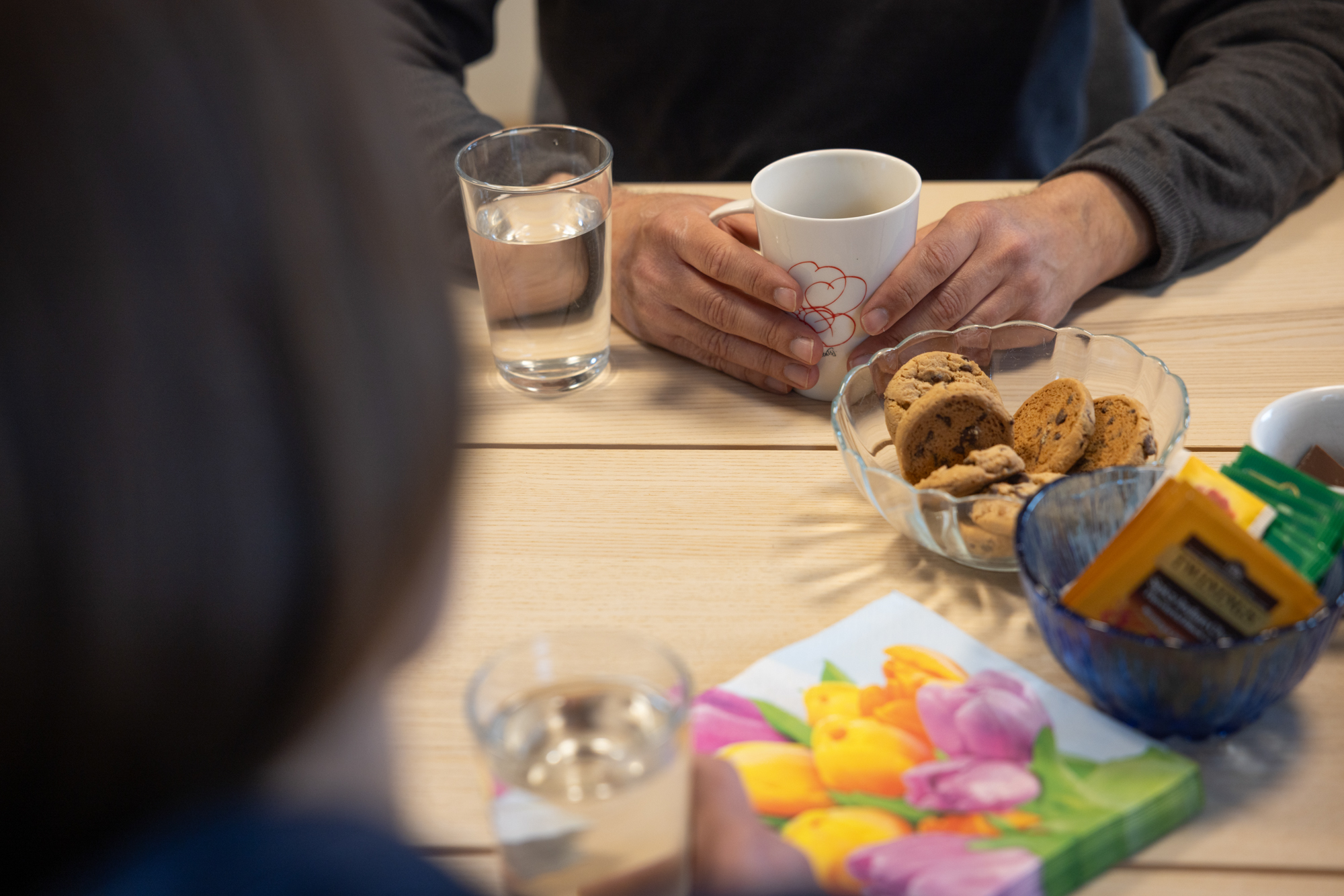 Foto: Nærbilde av et bord hvor det er satt fram kjeks, teposer og servietter. I forgrunnen sees et bakhode uskarpt, på motsatt side av bordet sees hendene til en mann som holder rundt en kopp.