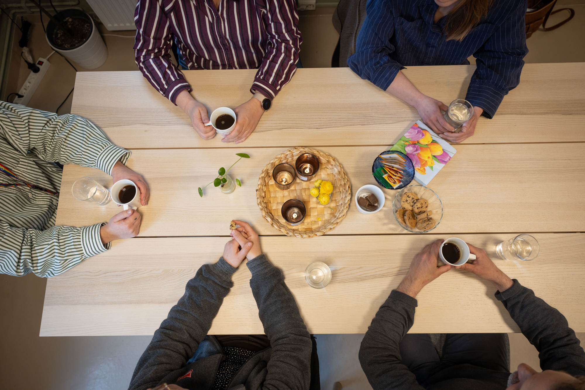 Foto: bilde tatt ovenfra av fem personer som sitter rundt et trebord. Bare armer og hender synes. personene holder i kaffekopper eller vannglass, og midt på bordet står te, servietter og kjeks.