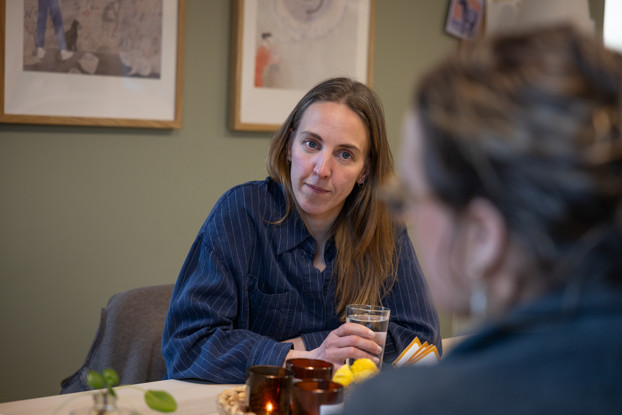 Foto: En kvinne med langt, brunt hår og stripete blå skjorte, sitter ved et bord og holder i et vannglass. Hun har et alvorlig uttrykk i ansiktet og ser mot en kvinne som sitter med ryggen til kamera, vendt mot henne. Kvinnen er uskarp i forgrunnen av bildet.